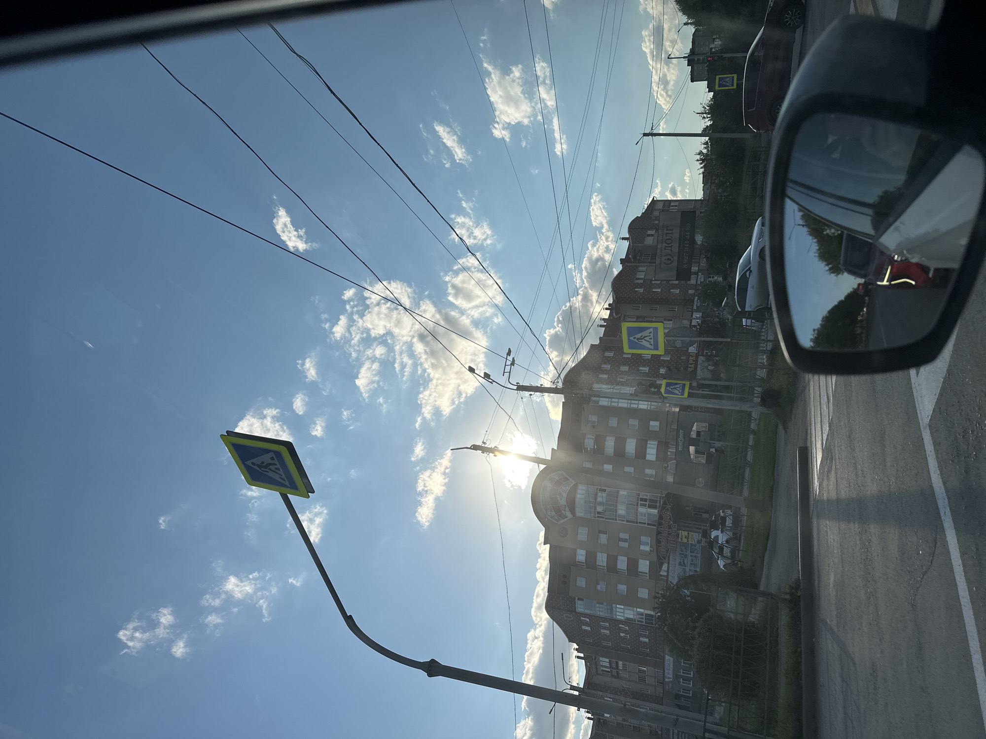 The image depicts a street scene in the city of Uchaly, Russia, as viewed from inside a vehicle. The sky is blue with scattered white clouds. Several power lines stretch across the sky. A pedestrian crossing sign, a blue square with a white depiction of a person walking, is visible on a pole. In the background, there is a multi-story brick building with numerous windows. Sunlight glints off the building and the sky. The road in the foreground is asphalt with white lane markings. A side-view mirror of the car shows reflections of other vehicles and trees, indicating movement or being stopped in traffic. Several other cars are visible on the road, including a silver SUV and a red crossover. Another pedestrian crossing sign is present further down the street. The overall impression is an urban environment during daylight hours.