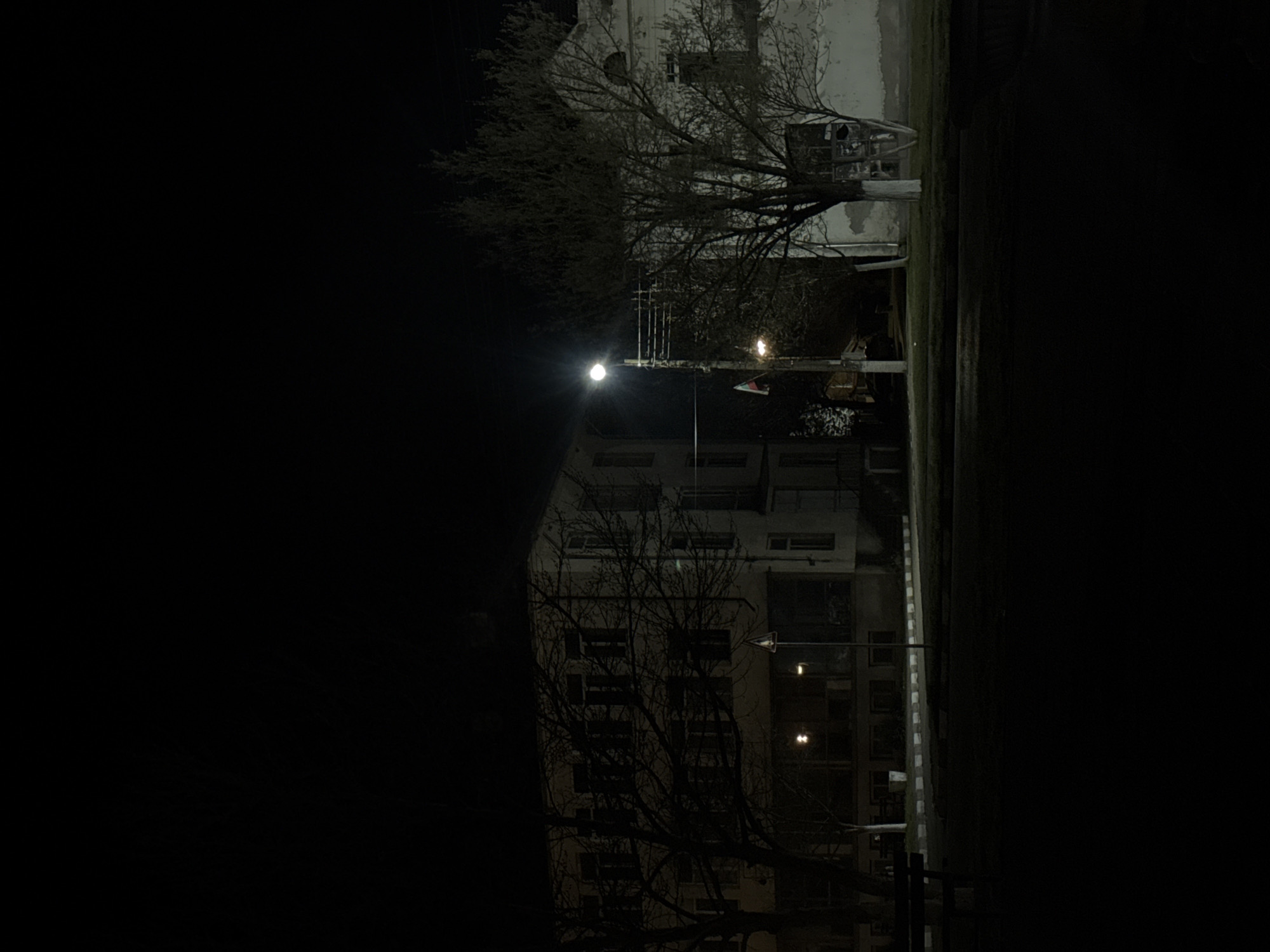 The image shows a night view in Lehchevo, Bulgaria. A multi-story building is partially illuminated by streetlights. Bare trees are visible in front of the building. To the right of the building, there appears to be a narrow strip of land. The rest of the image is dark.