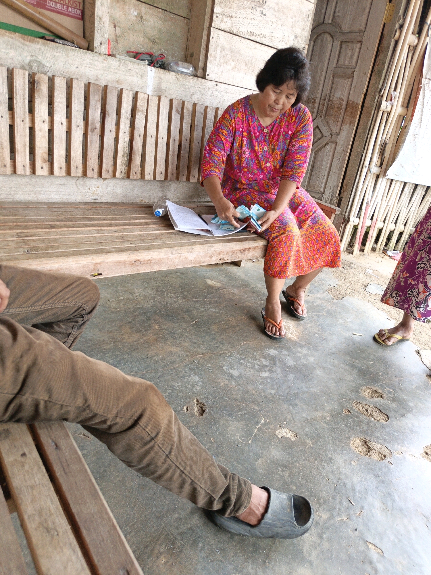 In Gedung Nyapah, Indonesia, a woman in a vibrant pink and orange patterned dress is seated on a wooden bench. She is holding what appears to be cash and documents. To her right, another person's legs are visible, wearing a patterned skirt and yellow flip-flops. In the foreground, a person wearing brown corduroy pants and dark grey clogs is partially visible, with their legs resting on the bench. The environment appears to be an outdoor, rustic setting with a concrete floor and wooden structures.