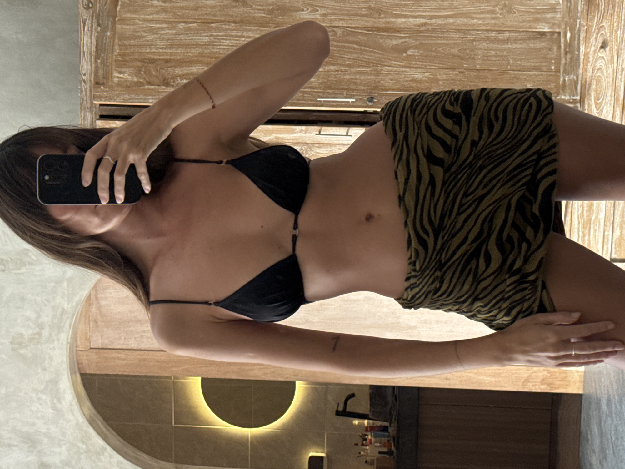A woman takes a mirror selfie inside a bathroom. She is wearing a black bikini top and a short olive green skirt with a zebra stripe pattern. She holds a black smartphone in her left hand. The bathroom contains a wooden vanity cabinet above her and a curved alcove sink with a yellow LED light behind it. The photo was likely taken in North Kuta, Indonesia.