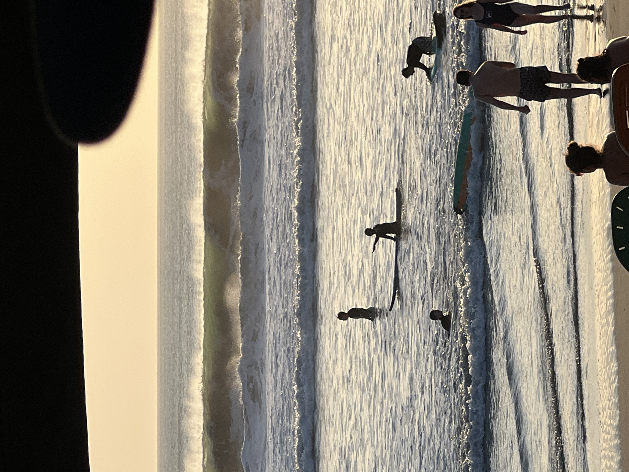 The image depicts a beach scene in Kuta, Indonesia, during the golden hour. Several individuals are present in the shallow water and on the sand. In the water, a few people are on surfboards, some paddling, and others standing. One person is partially submerged in the water. On the beach, two individuals are walking towards the water, and two more are seated in chairs in the foreground. The ocean displays moderate waves breaking, with a larger wave forming in the background. The sky is clear and illuminated by the setting sun, casting a warm glow over the scene.