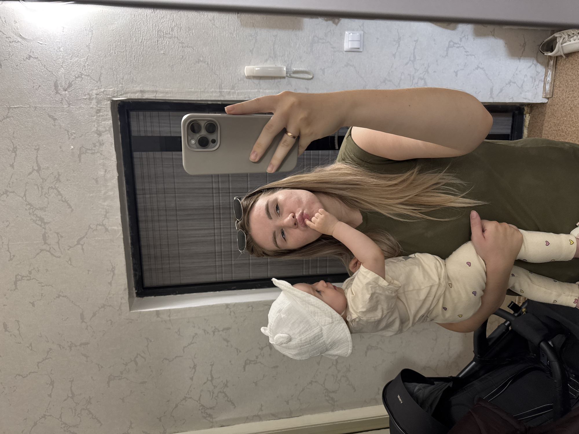 A woman is holding a baby and taking a selfie indoors. The woman has blonde hair and is wearing a green shirt. The baby is wearing a white outfit and a white hat with ears. The woman is holding her phone up to take the picture. There is a window behind the woman, and a stroller to the right. The background shows a wall with a switch and an intercom phone.
While it is possible the photo was taken in Chaikovsky, Russia, this cannot be conclusively determined from the image. There is no visible information to suggest the photo was taken in the city of Chaikovsky.