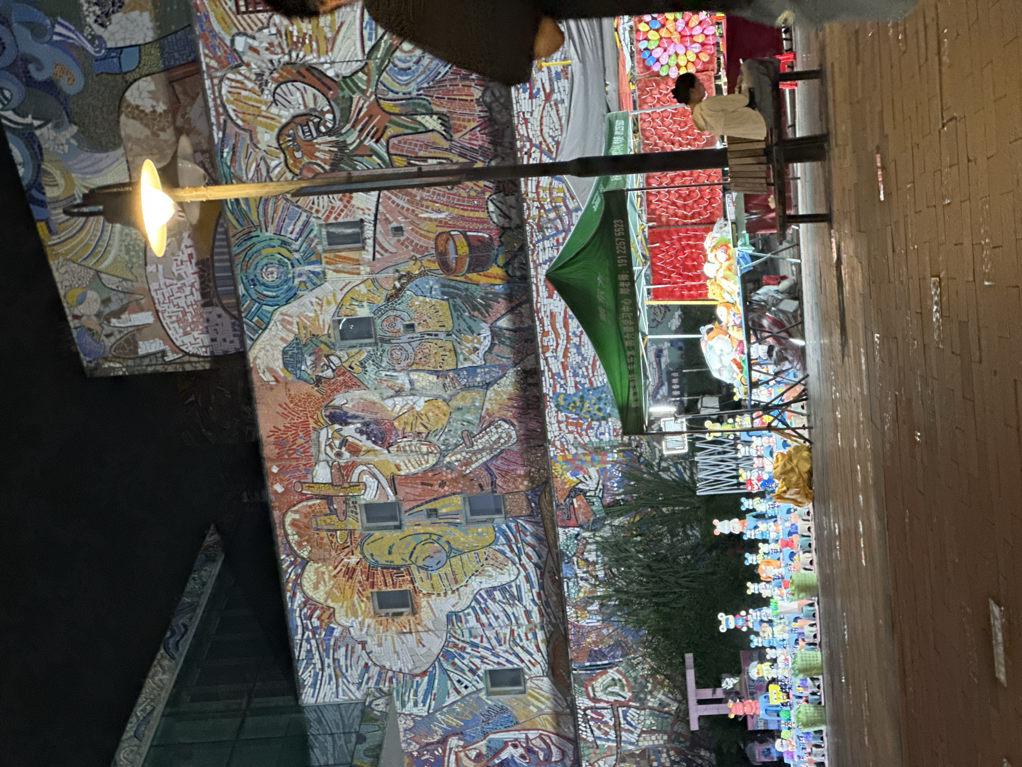 A street scene at night in the Chongqing High-tech Zone, China, features a large, vibrant mosaic mural on a building facade. In the foreground, a marketplace is set up with various stalls. One stall displays an assortment of colorful stuffed animals and toys arranged on tables and shelves. A young woman sits on a wooden bench, looking at her phone, in front of another stall selling what appear to be balloons. A street lamp with a glowing bulb stands near the center of the frame, illuminating the area. The ground is paved with bricks.