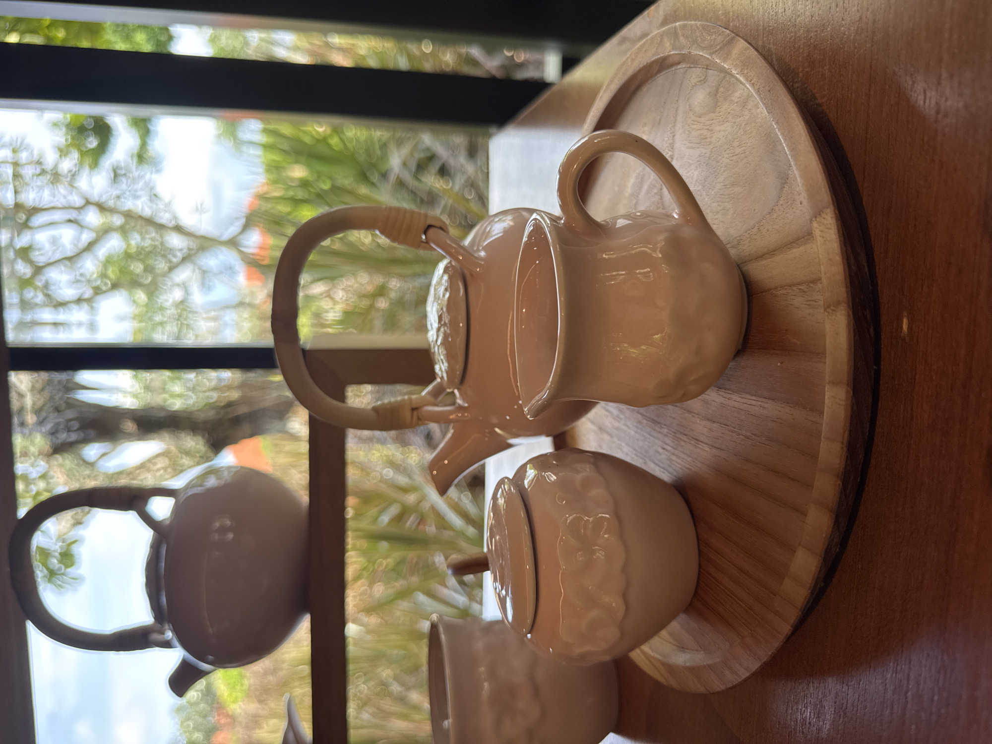 A tea set, including a teapot with a cane handle, a creamer, and a sugar bowl with lid, sits on a round wooden tray on a wooden table. The set and tray are in a peach color. In the background, a window shows a blurred exterior scene with greenery. The location is Kuta Selatan, Indonesia.