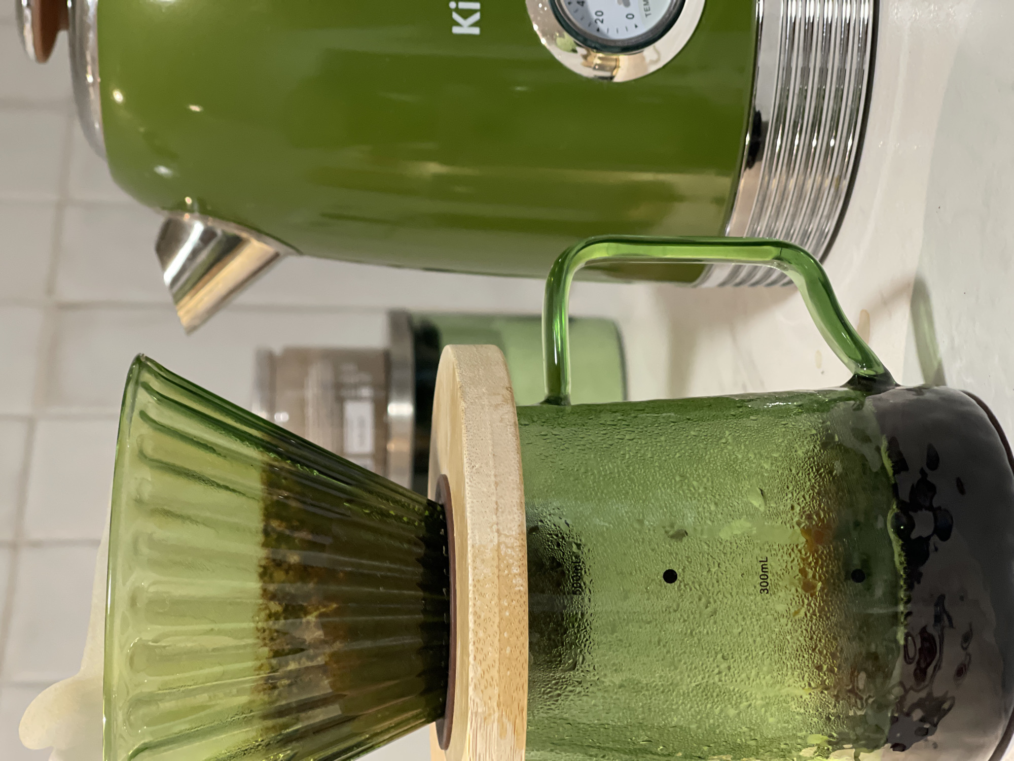 The image shows a close-up of a green kettle with silver accents and a matching green glass pour-over coffee maker. The coffee maker has a bamboo collar and a dark green bottom section. Condensation is visible on the glass. The kettle appears to be the brand "K". The background is a white tiled surface. The location and city are not discernible from the image.