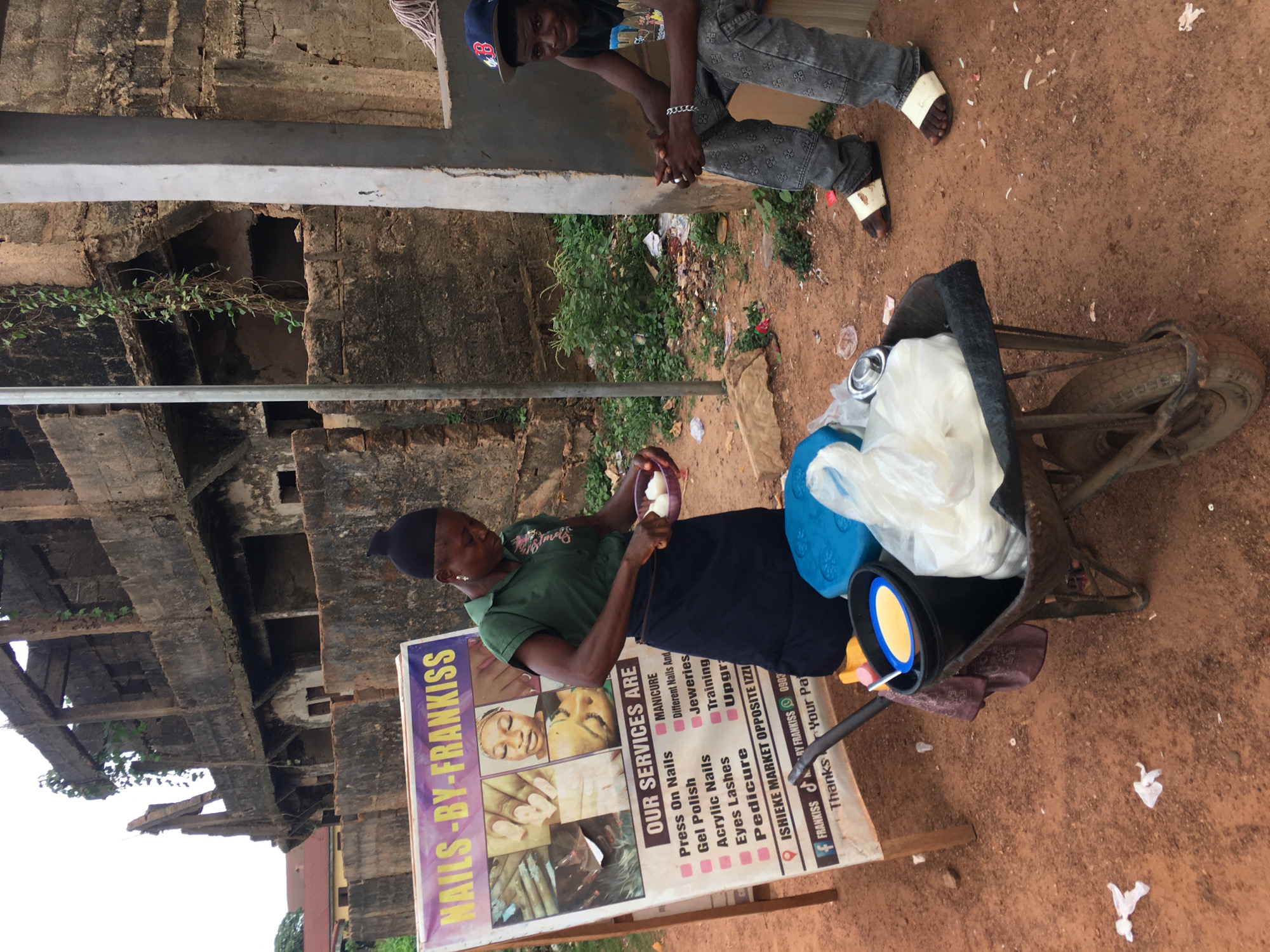 This image captures a lively street scene in Isieke, Nigeria, showing two individuals and a mobile nail salon setup. The setting is outdoors, characterized by reddish-brown earth and a weathered, multi-story concrete and brick structure in the background, partially overgrown with green vines, suggesting an older building or perhaps one under construction/deconstruction.
In the foreground, a woman, dressed in a green polo shirt and dark skirt with a black head covering, is seated on what appears to be a modified wheelbarrow or cart. She holds a small bowl containing white contents, seemingly engaged in an activity related to her nail services or possibly preparing food. Beside her, a large banner stands prominently, advertising "NAILS-BY-FRANKISS." The sign lists "OUR SERVICES ARE" Manicure, Press On Nails, Gel Polish, Different Nails, Acrylic Nails, Jeweleries, Eyes Lashes, Training, Pedicure, and Upgrade. It also includes contact information with social media handles and the address "ISHIEKE MARKET OPPOSITE IZZI," explicitly confirming the location.
Above and slightly behind the woman, a young man wearing a cap, a graphic t-shirt, and patterned jeans sits on a concrete ledge. He looks down towards the woman and the cart with a slight smile, his hands clasped. The ground around the cart shows some discarded items, typical of a bustling market area. The time of day is daytime, indicated by the natural light, and the overall atmosphere appears active and informal.