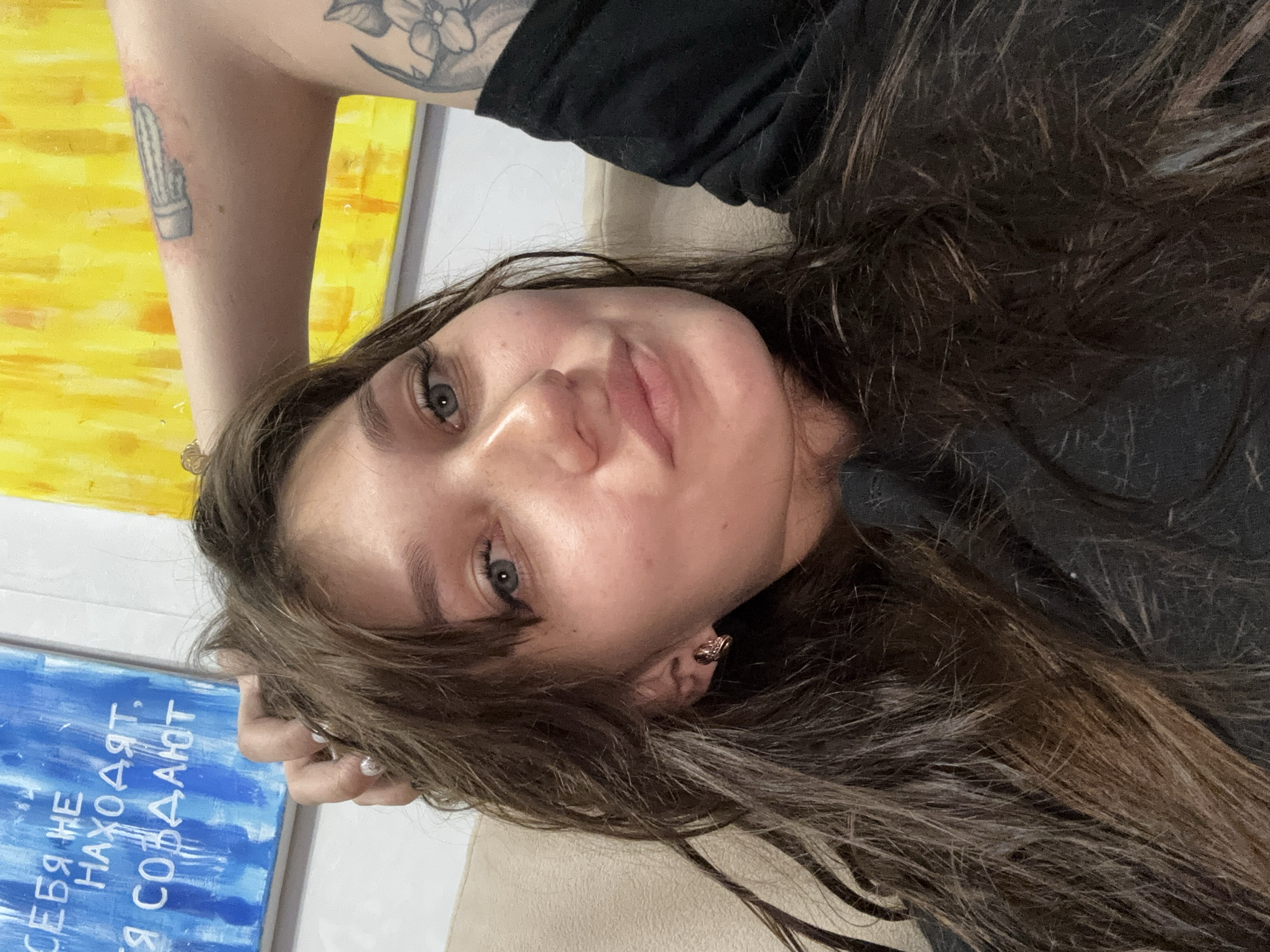 A woman with long, dark brown hair and blue eyes is lying on a couch, her head resting on her arm. She is wearing a black t-shirt. Behind her, a painting with blue and yellow abstract art is visible. The painting contains white text. A tattoo is visible on her upper arm, showing a flower. The scene appears to be indoors, possibly a living room. The location is not discernible from the image itself; there is no information indicating it is in Novokuznetsk, Russia.