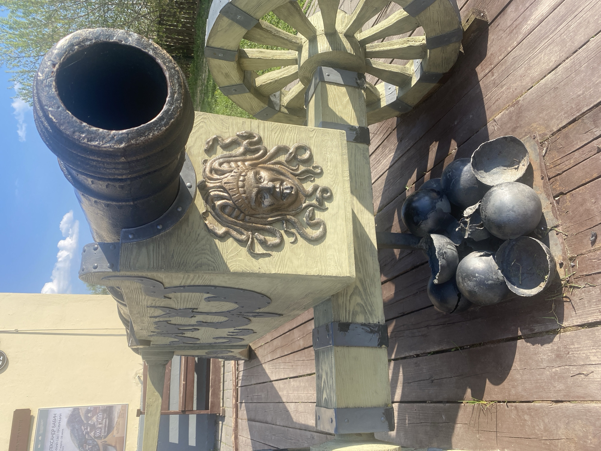 A stationary cannon installation sits on a wooden platform in Sviyazhsk, Russia. The cannon's barrel, made of dark metal, is positioned to the left. The carriage of the cannon, constructed of light wood, features a relief carving of Medusa. The cannon's wheels are large and wooden. To the right of the cannon, a metal tray holds cannonballs, some of which are broken open. The platform is made of wooden planks. In the background is a light-colored building.