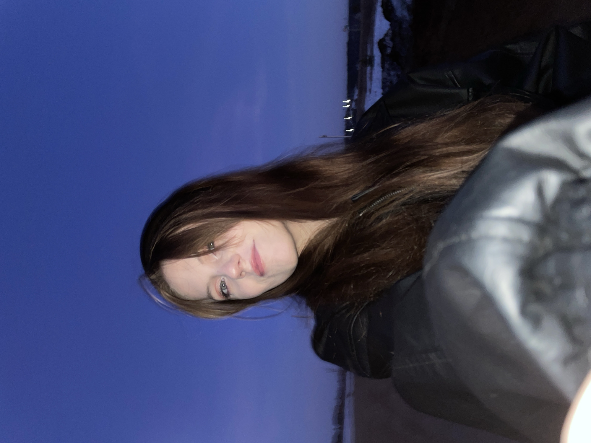 A rotated, close-up, eye-level outdoor shot shows a young woman with long brown hair wearing a black jacket. The background features a blue sky and a distant, indistinct cityscape, possibly identifying the location as Фоки, Russia. The woman is looking at the camera. The lighting is dim, suggesting dusk or nighttime.
