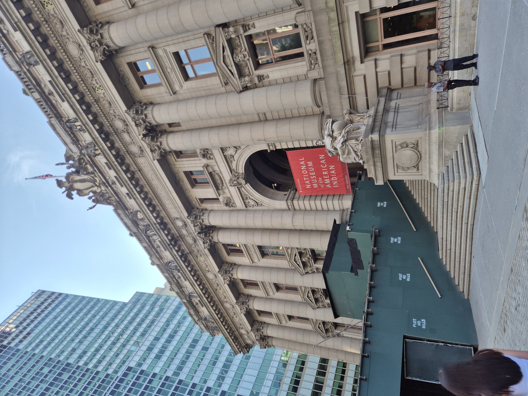 The image captures the National Museum of the American Indian, located in the U.S. Custom House at One Bowling Green in the Financial District of New York City. The grand, classical facade of the building is the primary subject, characterized by its large Corinthian columns, ornate carvings, and numerous windows. A prominent red banner hangs from the facade, bearing the text "Smithsonian NATIONAL MUSEUM of the AMERICAN INDIAN" and "FREE ADMISSION."
In front of the building, a statue of a seated figure with a globe stands on a pedestal. To the left of the main entrance, construction fencing, painted a dark green, surrounds a section of the building, with "POST NO BILLS" stenciled in white letters in several places. A set of wide stone stairs leads up to the entrance, partially obscured by the construction.
Two men, possibly security guards, are standing on the sidewalk in front of the building. One is wearing a dark uniform shirt and pants, the other a dark t-shirt and pants. A woman with dark hair, seen from behind, is walking towards the left edge of the frame in the foreground.
In the background, a modern skyscraper with a glass facade rises to the left of the historic building, creating a contrast between old and new architecture. The sky above is a clear light blue. The overall scene depicts a public space in a bustling urban environment, with elements of historical preservation and ongoing construction.