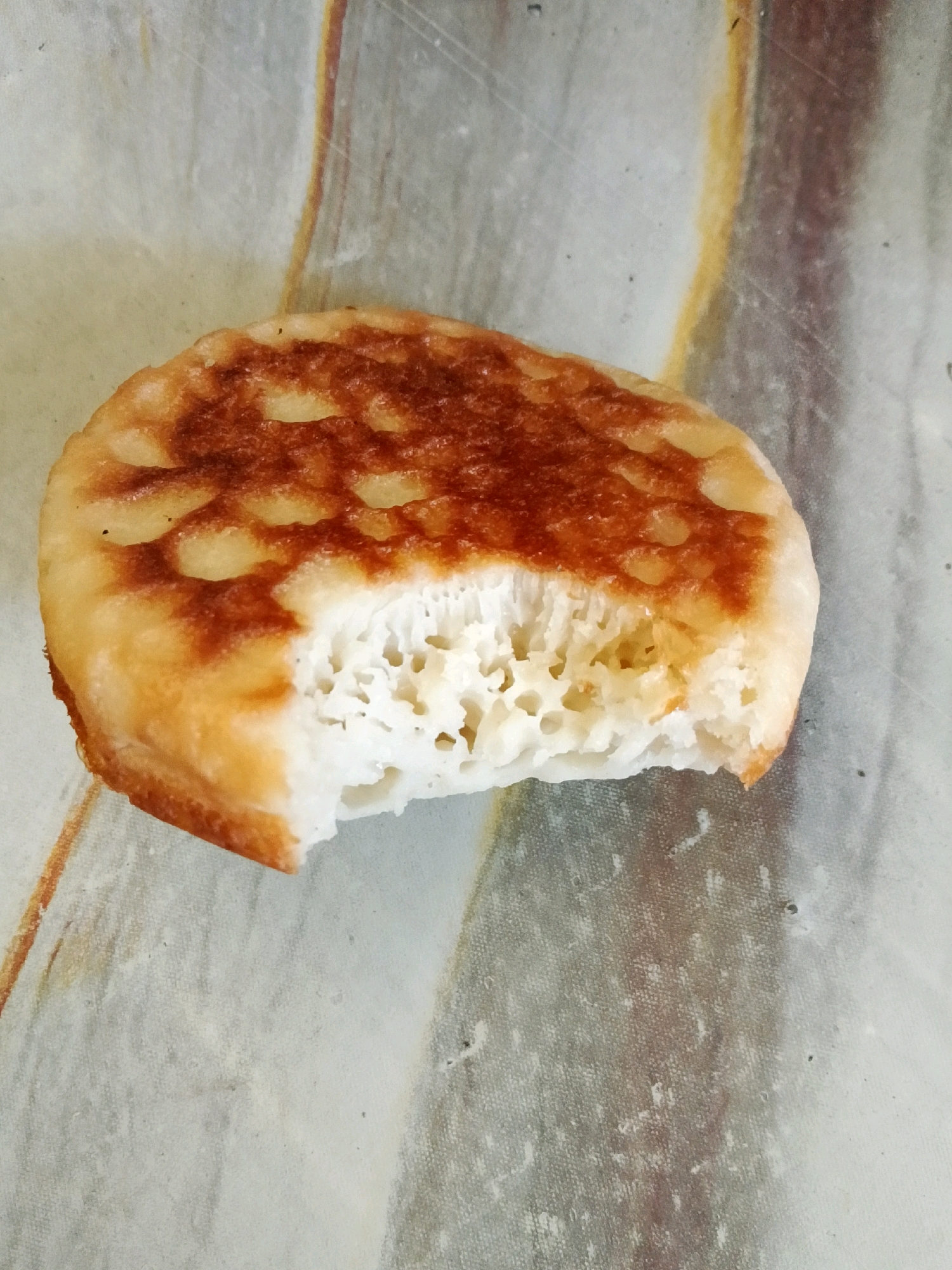 A single, round, golden-brown pancake has a bite taken out of it. The pancake rests on a light-colored surface with a marbled pattern of brown and white. The pancake's top is pockmarked with small, circular indentations, and its interior is airy and white with visible holes. The background surface appears textured and possibly slightly worn. There are no people or any other objects visible in the image. The image does not provide any information about a specific location such as the city of Kalikondang, Indonesia.
