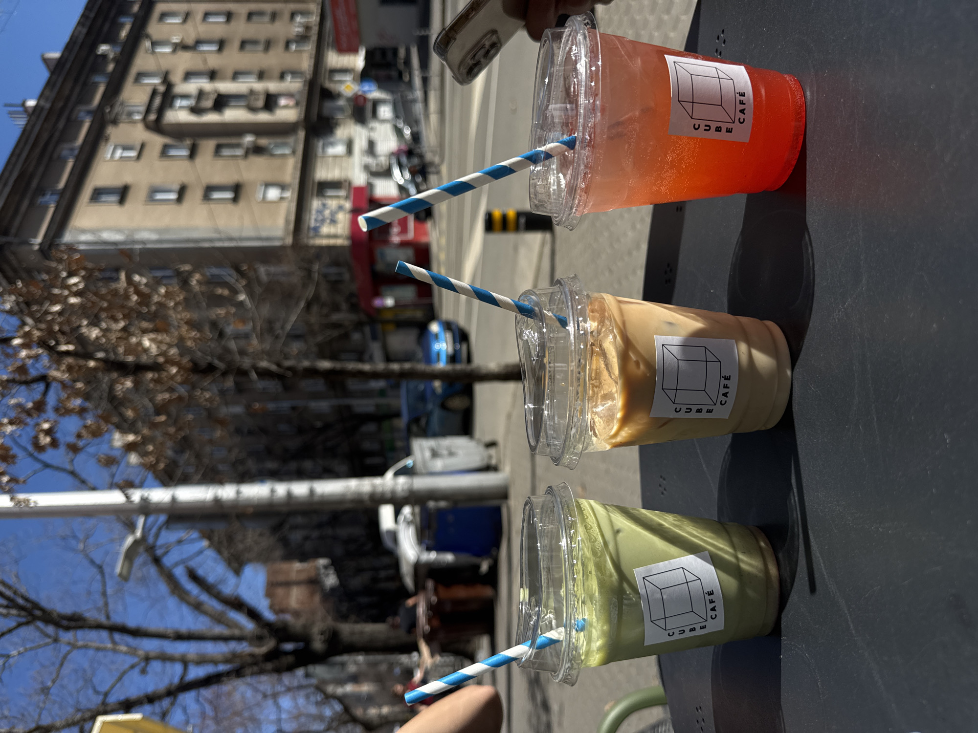 The image shows three plastic cups with blue-and-white striped straws, sitting on a dark grey table. Each cup contains a different drink: the first one appears green, the second one is coffee-colored, and the third one is orange. Each cup has a white sticker with the word "CUBE" and "CAFÉ" next to a cube-shaped drawing. The background shows an urban scene. Buildings are visible, along with trees and cars. A red truck is also noticeable. The location is likely Sofia, Bulgaria.