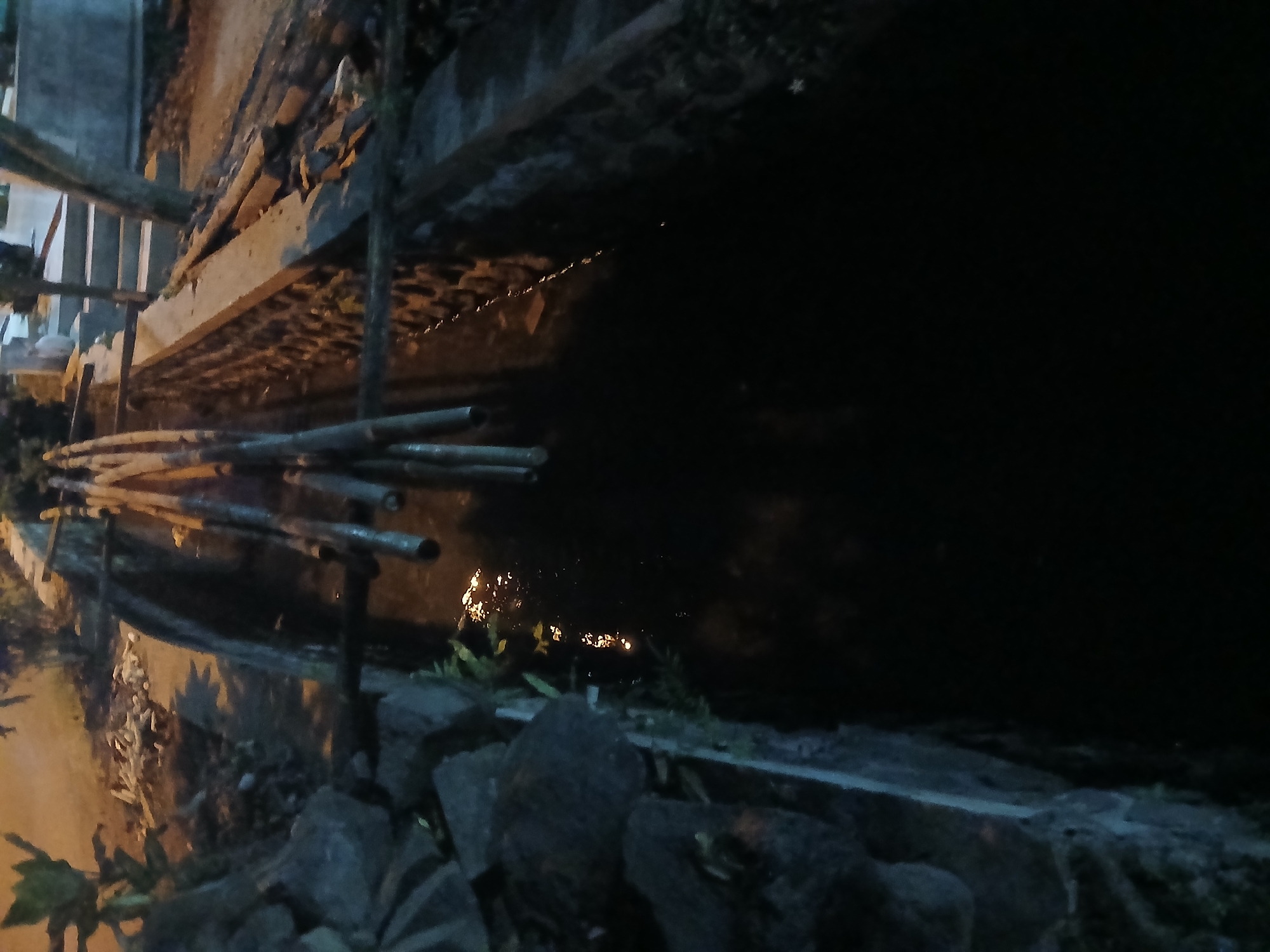 The image shows a narrow, dark channel of water, likely a ditch or canal, running through an outdoor area. The sides of the channel are lined with rough stone and brickwork. Several long, thin wooden poles are placed across the channel. The water is dark and reflects some light, suggesting it is dirty or stagnant. The surrounding environment appears to be a mix of natural elements, such as rocks and plants, and man-made structures, including steps and possibly a pathway or road in the background. The lighting indicates it is either dusk or nighttime. There are no people or animals visible in the image. This scene is located in Kademangan Kulon, Indonesia.