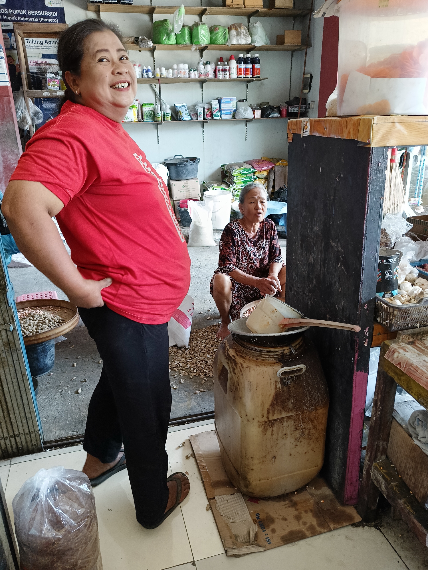 The image depicts an indoor scene, likely a shop or market stall, in Manisrenggo, Indonesia. In the foreground, a woman in a red t-shirt and black pants stands facing left, with her left hand on her hip. She is smiling and looking to her right. To her left is a woven basket filled with small, light-colored objects. Behind her, a second woman, older and wearing a patterned dress, is crouching and looking forward, with her hands clasped in front of her. She is positioned near a large, opaque plastic container that appears to be a jug or drum, which is sitting on a piece of cardboard. The background reveals shelves stocked with various items, including bags of what appears to be fertilizer or feed, bottles, and other goods. The floor is tiled, and there are scattered debris and discarded materials around the area. The overall impression is of a functional, perhaps slightly cluttered, commercial space.