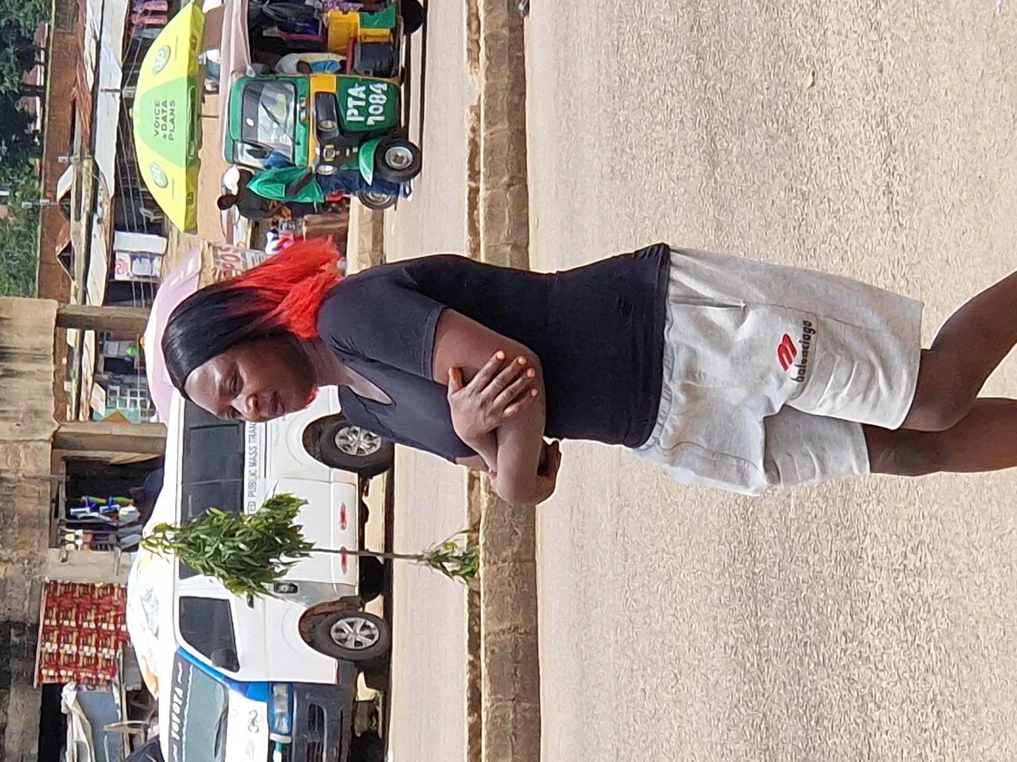 A medium shot captures a Black woman with vibrant red-tipped black hair, wearing a black t-shirt and light gray shorts, standing in a street. Her arms are crossed, and she is facing to her left. Behind her, a white van with the word "TOYOTA" visible is parked. Further in the background, there are market stalls with various goods displayed and a colorful auto-rickshaw. The scene appears to be a busy street in Kabong, Nigeria, with elements suggesting a bustling market environment. The ground is paved, and a low stone barrier runs along the side of the road. The lighting suggests it is daytime.