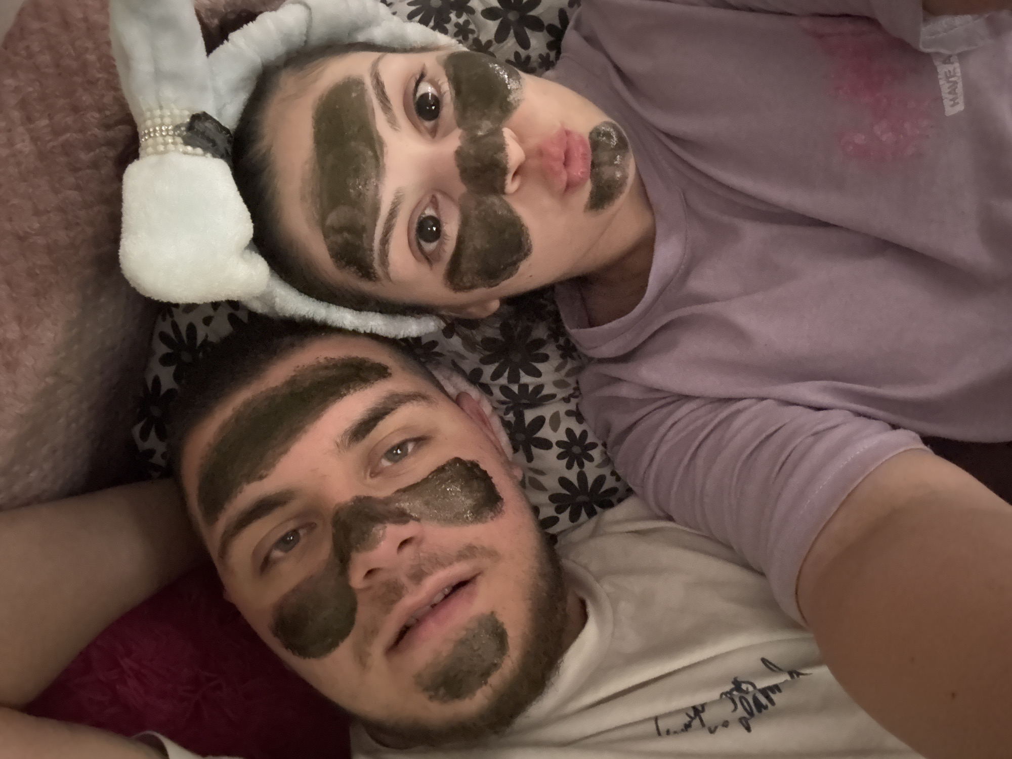 The image captures a high-angle self-portrait of two individuals, a male and a female, lying closely together. Both subjects have patches of a dark green or black facial mask applied to their faces.
The male, situated on the left, has short dark hair, light-colored eyes, and visible stubble on his chin and jawline. Dark mask patches are present on his forehead in stripe-like patterns, under his left eye, on his right cheek, and above his upper lip. He is wearing a white t-shirt with dark script visible on the left side of the chest. His expression is neutral with slightly parted lips.
The female, positioned on the right, has dark hair held back by a light blue or grey fluffy headband adorned with a prominent white bow embellished with pearls and rhinestones. Mask patches are applied to her forehead, under both eyes, on her nose, cheeks, and chin. She is wearing a light purple long-sleeved t-shirt with a faint pink design and partially visible white text "HAVE A" on the lower right front. Her lips are pursed in a pout, and her eyes are wide, directed towards the camera.
Both subjects are resting on a dark grey pillow with a white floral pattern. Behind the male's head, a dark red fabric is visible, and a light pink, fuzzy textile is in the upper background. The lighting is soft and ambient. There are no discernible visual elements within the image that specify the geographical location as Burgas, Bulgaria.