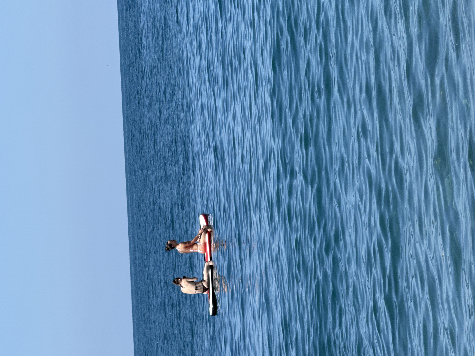 Two individuals are paddleboarding on a calm, blue sea under a clear sky. They are positioned on a single paddleboard, with one person in front and the other behind. The person in front is wearing a pink bikini and is looking to the right. The person behind is wearing a dark bikini and is looking to their left, holding a phone. The paddleboard is red and white. The scene is set in the city of Sarpi, Georgia.