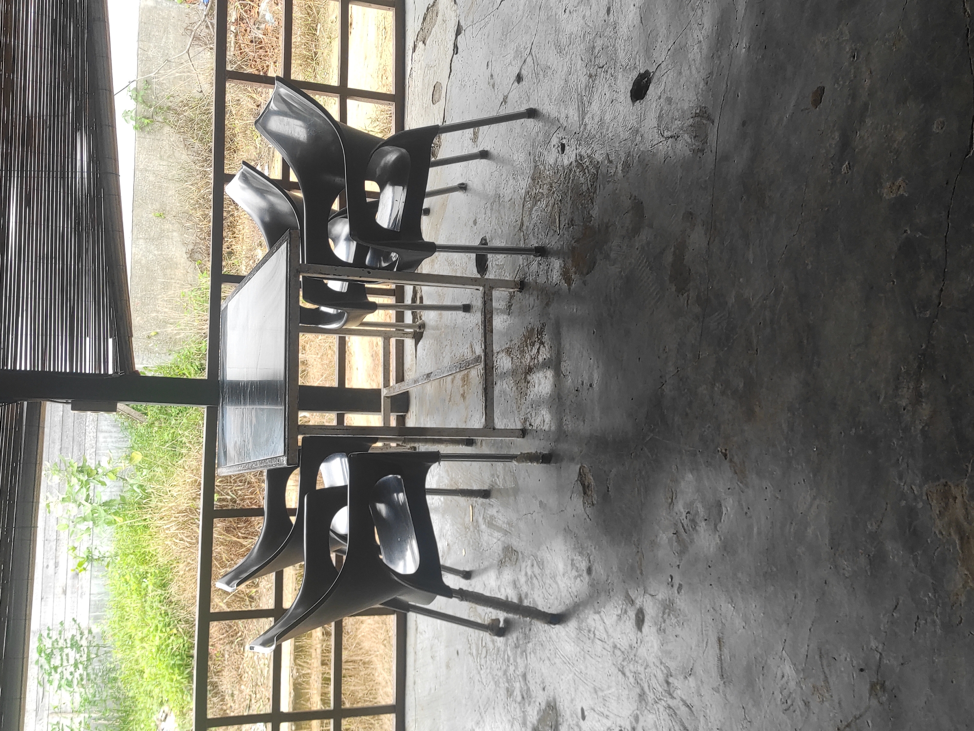 The image shows an outdoor seating area in Lhokseumawe, Indonesia. A metal-framed table is surrounded by four black plastic chairs. The floor is a worn, gray concrete surface with cracks and discoloration. A partially obscured view of vegetation is visible through a window with a metal grid. A bamboo curtain is visible at the far left of the image. There are no people visible.