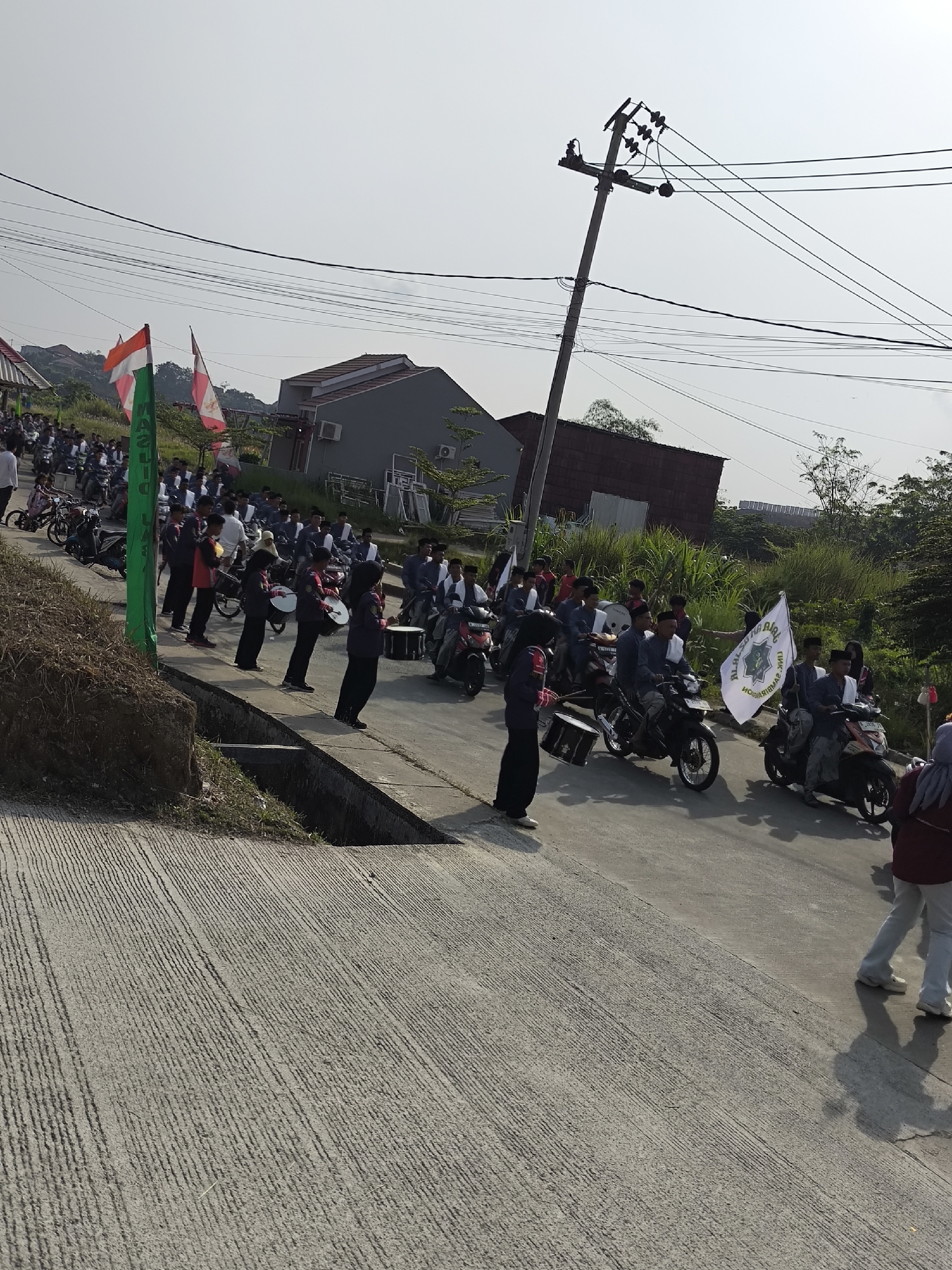 This is a photograph of a parade or procession taking place on a street in Cilegon, Indonesia. The scene is captured in daylight under a clear sky with some visible electrical wires and a utility pole dominating the upper portion of the image.
In the foreground, a paved road with distinct parallel grooves runs diagonally from the bottom left to the upper right. To the left of the road, there is a raised embankment covered with dry grass and vegetation. A concrete drainage channel runs along the edge of the road.
The main subjects of the photograph are a group of people, likely students, participating in a parade. Many of them are riding motorcycles, while others are walking. Several individuals are carrying musical instruments, specifically drums, and appear to be part of a marching band. Some people are holding flags; one is a red and white Indonesian flag, while another is a white flag with green text and a circular emblem. The participants are mostly dressed in dark uniforms, some with red accents, and many are wearing head coverings like peci hats or hijabs.
In the background, there are several residential buildings with dark roofs and light-colored walls, suggesting a suburban or semi-urban environment. Lush green vegetation and trees are visible behind the buildings. The overall impression is of an organized community event, possibly a cultural or celebratory parade, taking place on a sunny day in Cilegon.