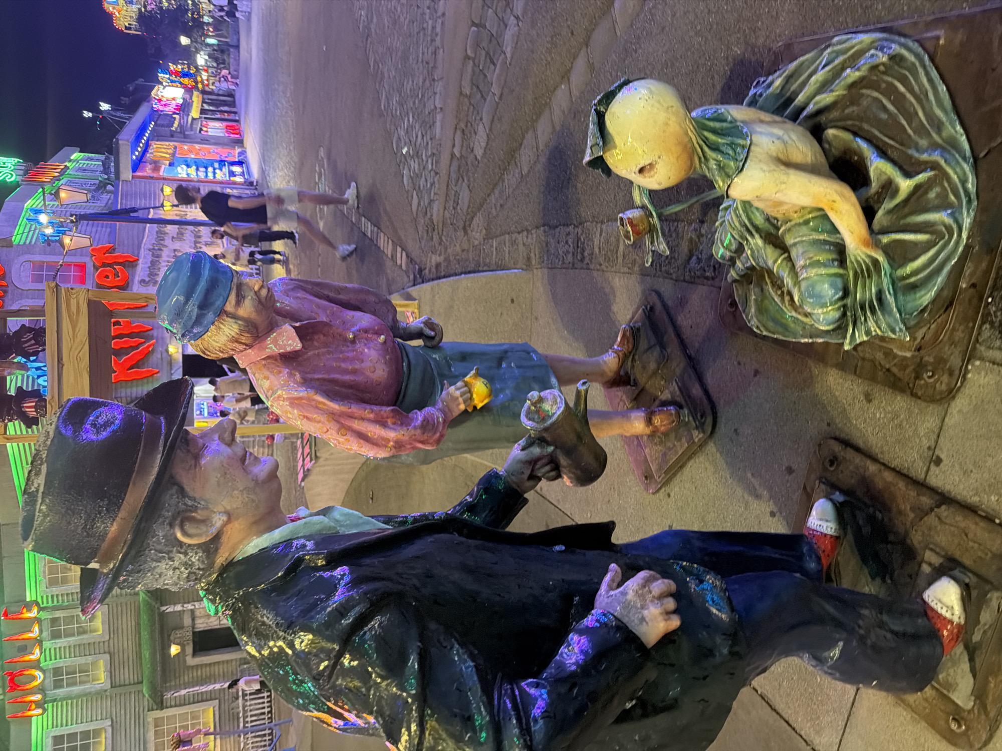 The image captures a nighttime scene on a city street, likely in Vienna, Austria, given the presence of illuminated neon signs typical of entertainment districts. In the foreground, two life-sized, weathered statues depict a man and a woman. The man, wearing a dark hat, a shiny black jacket, and blue pants, is holding a metal watering can, seemingly pouring its contents. The woman, with blonde hair under a blue cap and wearing a pink blouse and a blue skirt, is bent over, holding a small yellow object in her hand. In front of them sits another statue of a small, yellow, bulbous creature with a green garment, holding a tiny cup. The street is paved with cobblestones and lined with buildings adorned with vibrant neon lights, creating a festive and somewhat surreal atmosphere. Several real people can be seen in the background, walking along the street, blurred due to the low light conditions and movement. The overall impression is of a lively, possibly amusement-park-like, street setting with artistic sculptures.