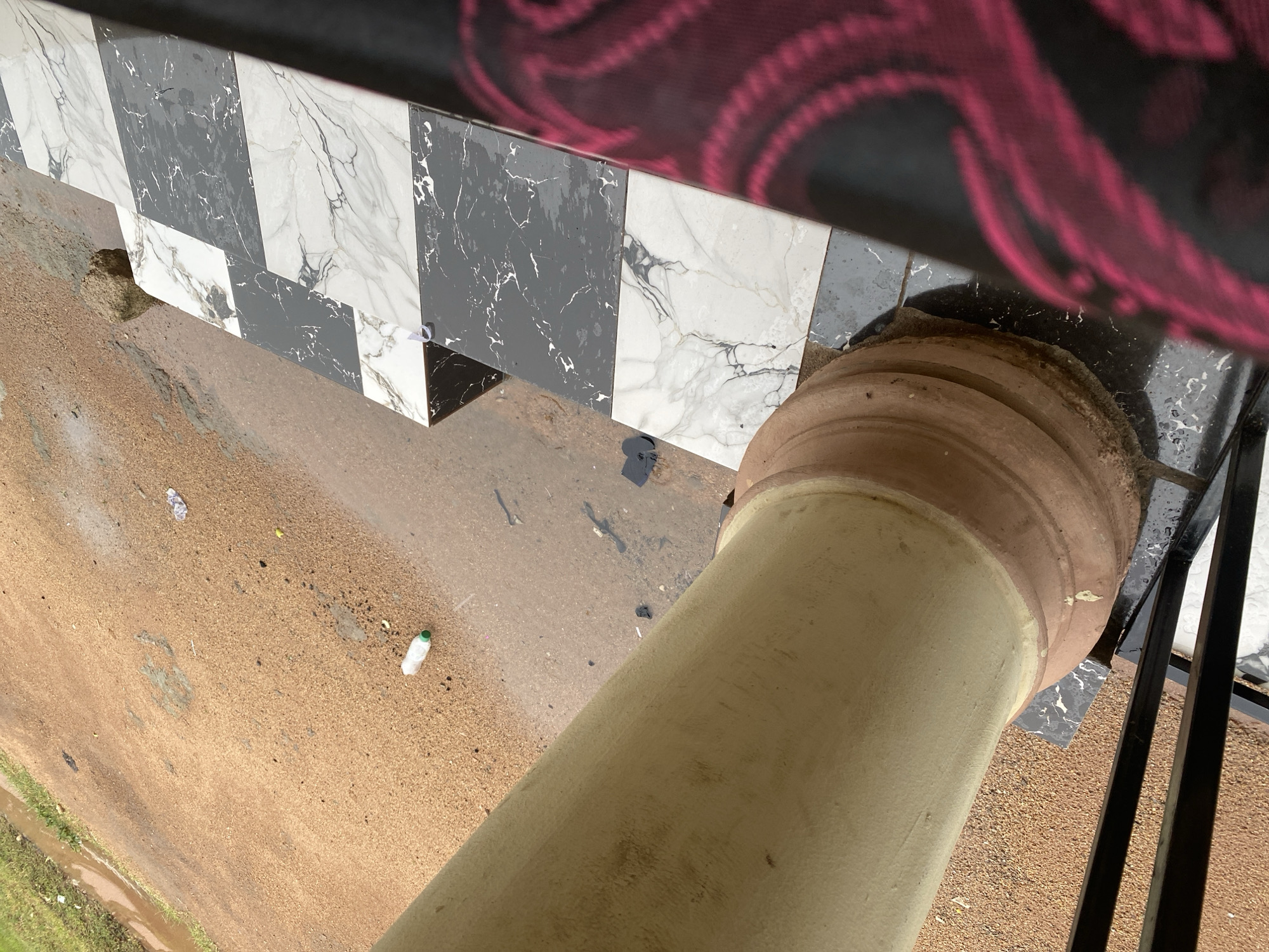 In Jalingo, Nigeria, an outdoor scene is depicted. A large, cream-colored pillar dominates the left side of the frame, showing some dirt and wear. To the right of the pillar, a tiled surface with a black and white marble pattern is visible, arranged in alternating rectangular blocks. A dark, patterned fabric, possibly clothing, is present on the far right edge of the image, featuring a design with pink or magenta swirls. The ground appears to be composed of reddish-brown earth, with patches of green grass along the top left edge. There are signs of recent rain, with puddles of water and dampness on the ground and tiles. Scattered on the ground are a few pieces of debris, including a white plastic bottle with a green cap, a crushed plastic bottle, and some dark, unidentifiable fragments. The overall impression is a somewhat neglected outdoor area, possibly a walkway or patio, with elements of both natural and man-made surroundings.