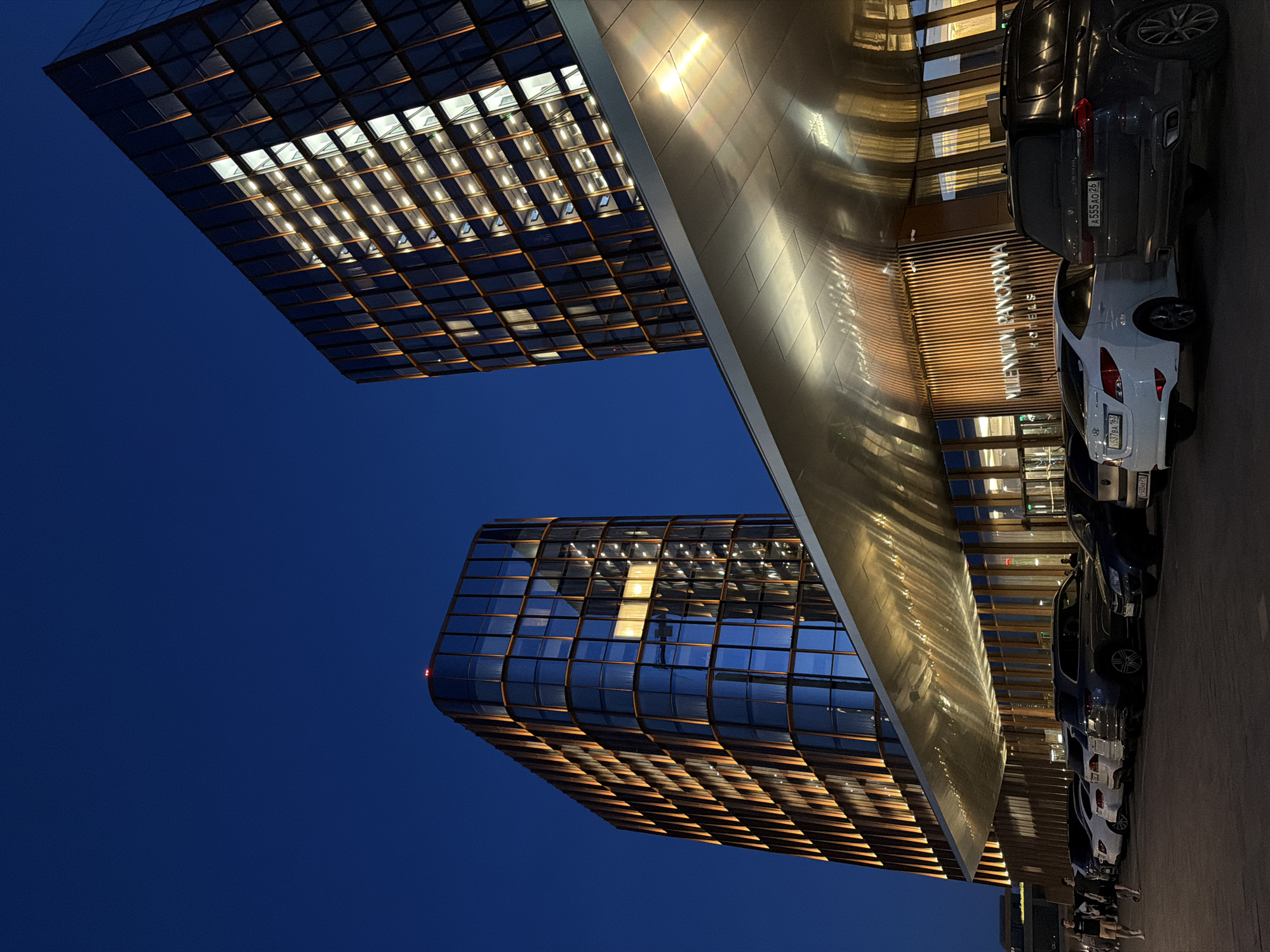 The image depicts the Millennium Panorama Hotel in Kazan, Russia, at night. The hotel building showcases modern architecture with large glass windows illuminated from within, creating a grid-like pattern. Several cars are parked along the side of the building. The sky is a dark blue. The image is taken from a low angle, accentuating the height of the buildings. The name "Millennium Panorama Hotels" is visible above the entrance.