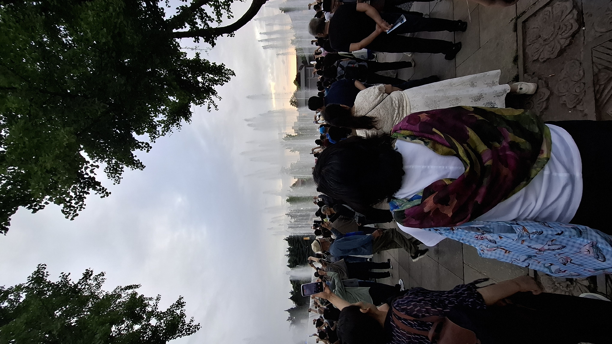 The image depicts a crowd of people gathered in Yanta District, China, likely at night. Many individuals are holding up their phones, presumably taking pictures or videos of a water fountain display. The fountains are arranged in a line, shooting water high into the air. Trees are visible in the foreground, framing the scene. The overall atmosphere suggests a public gathering for entertainment or sightseeing.