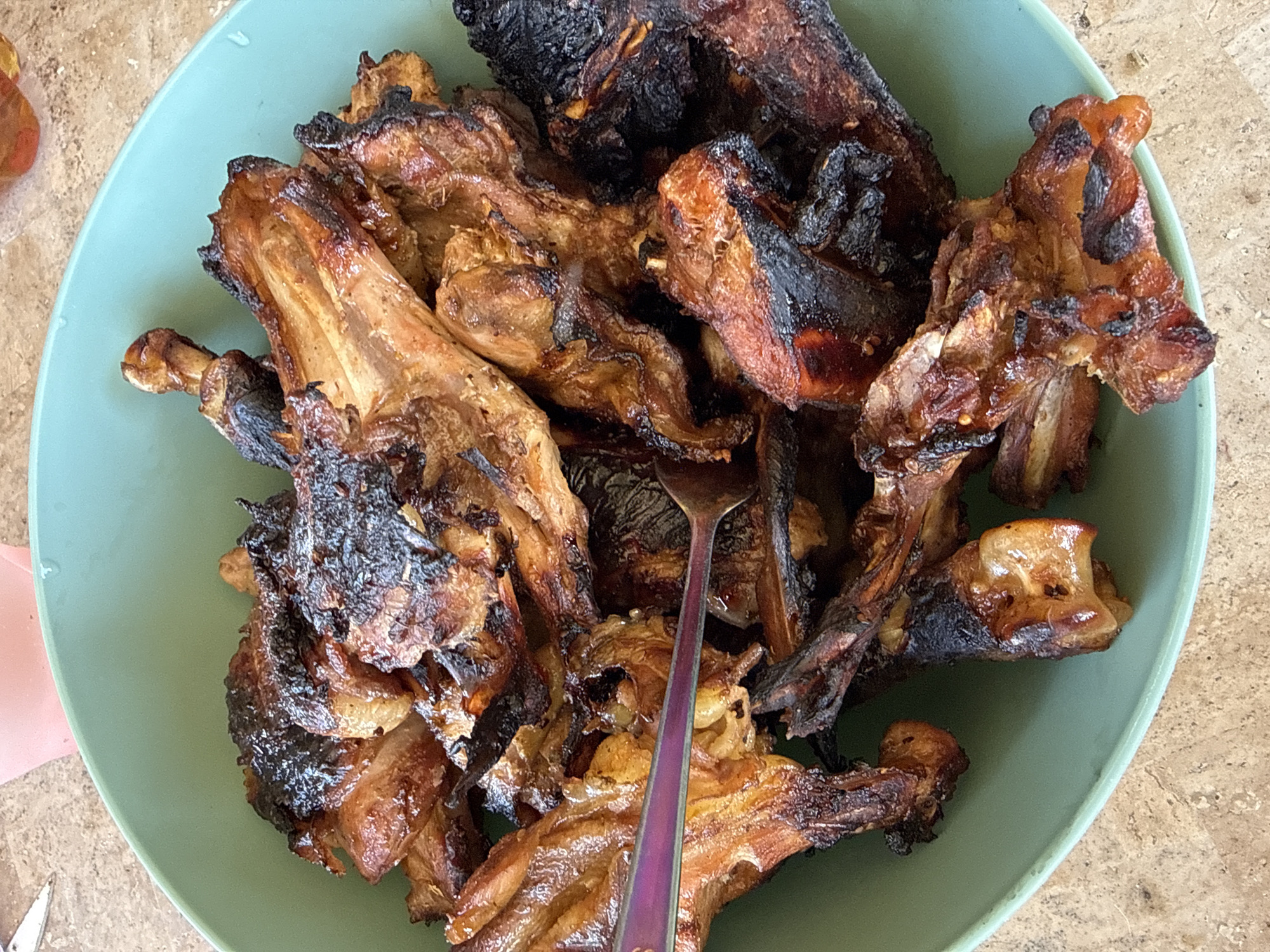 A close-up overhead shot shows a pile of cooked ribs in a teal bowl. The ribs are a deep brown and black, with some charring visible. A fork with a purple handle is stuck into the ribs, partially obscured by the meat. The bowl sits on a light brown, speckled surface that appears to be a countertop or table. In the background, a pink cup can be seen, slightly out of focus. The lighting suggests this image was taken indoors, possibly in a home kitchen in Horbanivka, Ukraine.