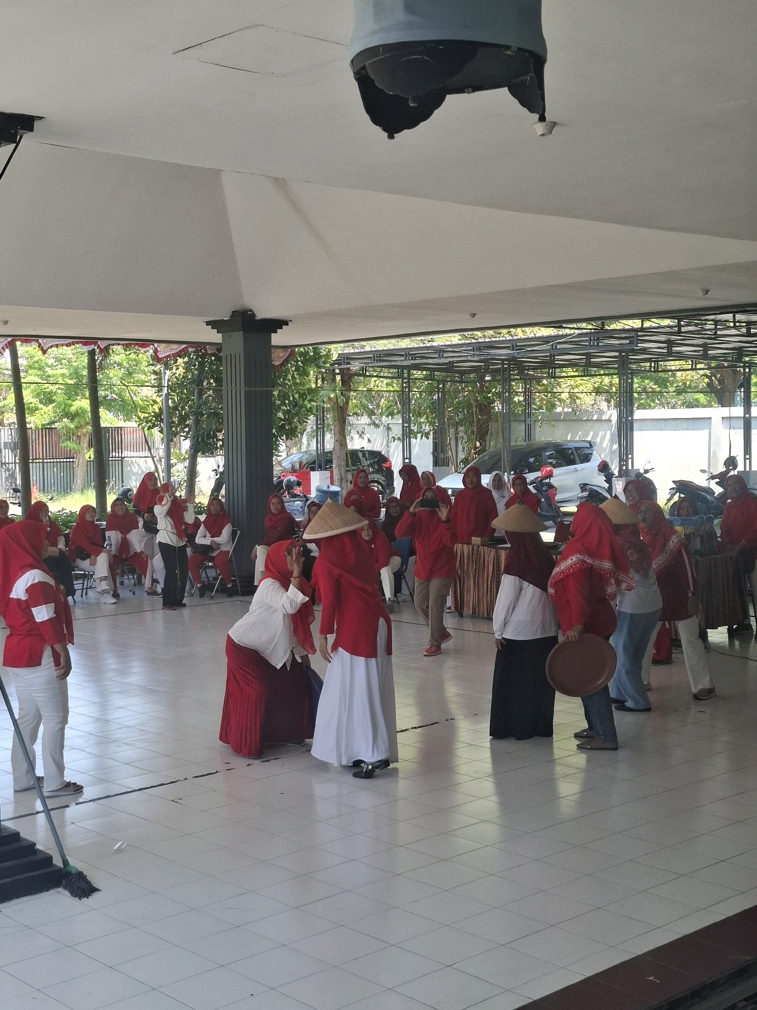 The image depicts an outdoor event with a group of people, mostly women, dressed in red and white attire, some wearing traditional conical hats. They appear to be engaged in some form of performance or activity in a tiled courtyard area. In the background, there are trees, a building with a white wall, a parked white car, and several motorcycles. The architecture suggests a public space with a covered seating or gathering area supported by pillars. The scene is likely taking place in Surabaya, Indonesia, given the presence of conical hats often associated with Indonesian culture and the general ambiance of the setting.