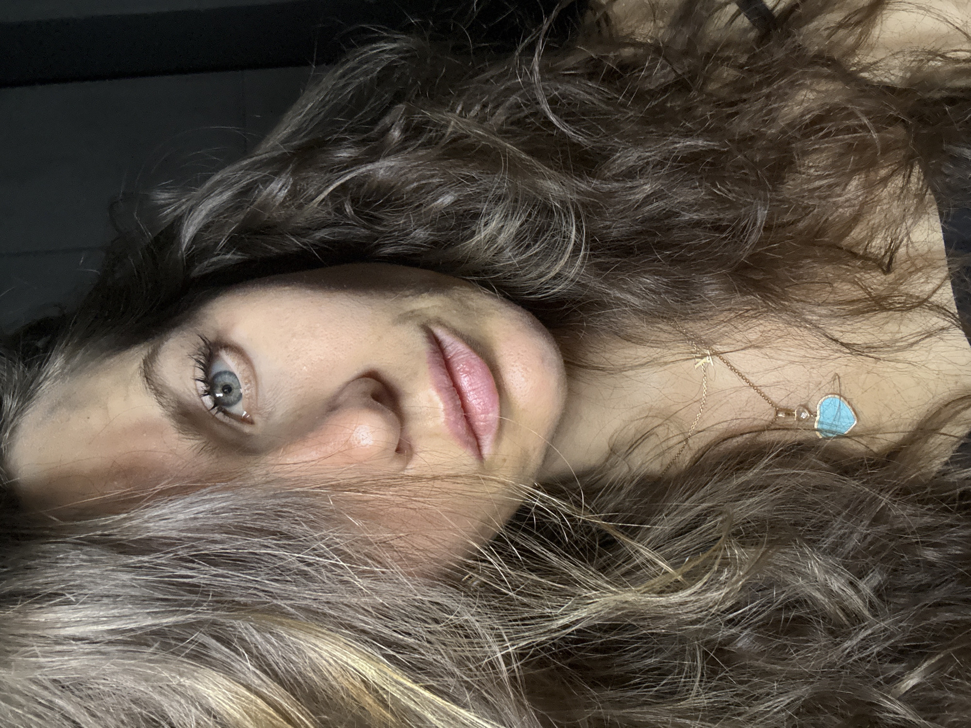 A close-up, rotated portrait captures a woman with long, wavy, multi-toned hair, appearing to lie down. Her head is positioned towards the left of the frame, with her body extending to the right. Her left eye, a striking blue, is open and looks directly towards the viewer. Her lips are softly parted, revealing a natural pink hue, and the bridge of her nose is visible.
Her hair, a blend of brown and lighter, possibly blonde or ash-blonde, strands, is spread out around her head, creating a textured frame for her face. Adorning her neck is a delicate gold-toned necklace, featuring a small "T" charm on its chain and a heart-shaped pendant crafted from a light blue stone, possibly turquoise, accompanied by a small, clear gemstone.
The lighting is soft, illuminating her face and the front of her hair from the right side, while the upper left corner of the image is dark and indistinct, suggesting a low-light environment or a dark background. Her expression is neutral and direct. No specific location cues for Khimki, Russia are visible within the image, nor is any specific activity other than posing for the photograph.