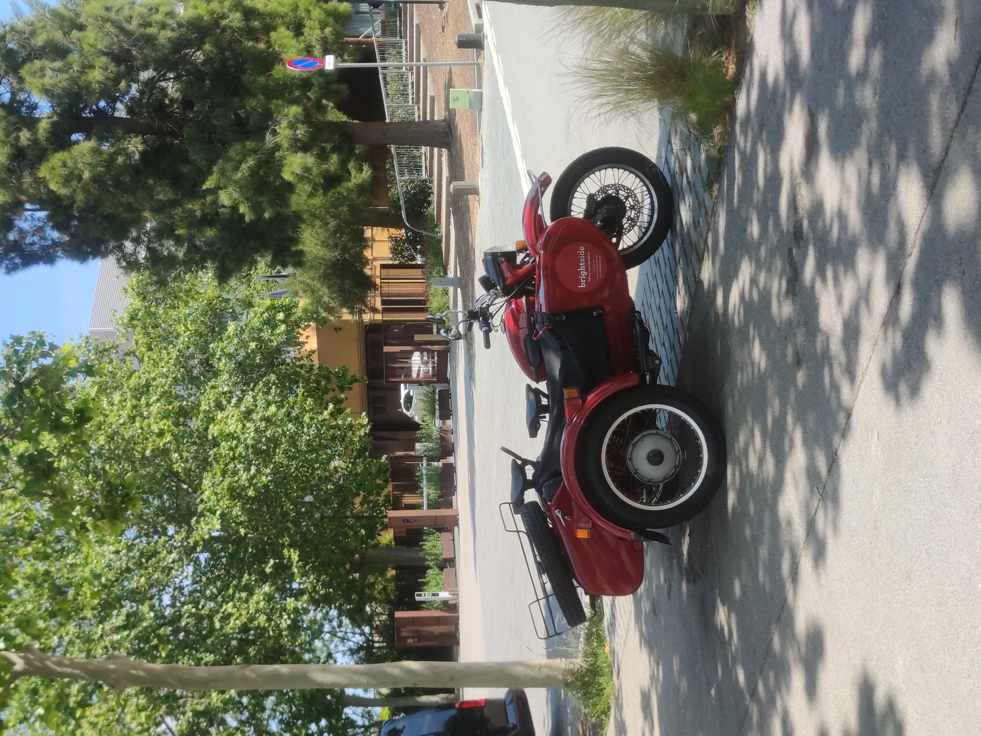 A red three-wheeled vehicle is parked on a sidewalk in an urban environment. The vehicle has the word "brightside" displayed on its side. The sidewalk is paved with gray concrete and is partially shaded by tree leaves. Trees and plants are visible along the side of the street, and a building is present in the background. A road sign is visible near the top center of the image. A partial view of a car is seen in the bottom-left corner. Based on the architectural style and vegetation, the location is in Barcelona, Spain.