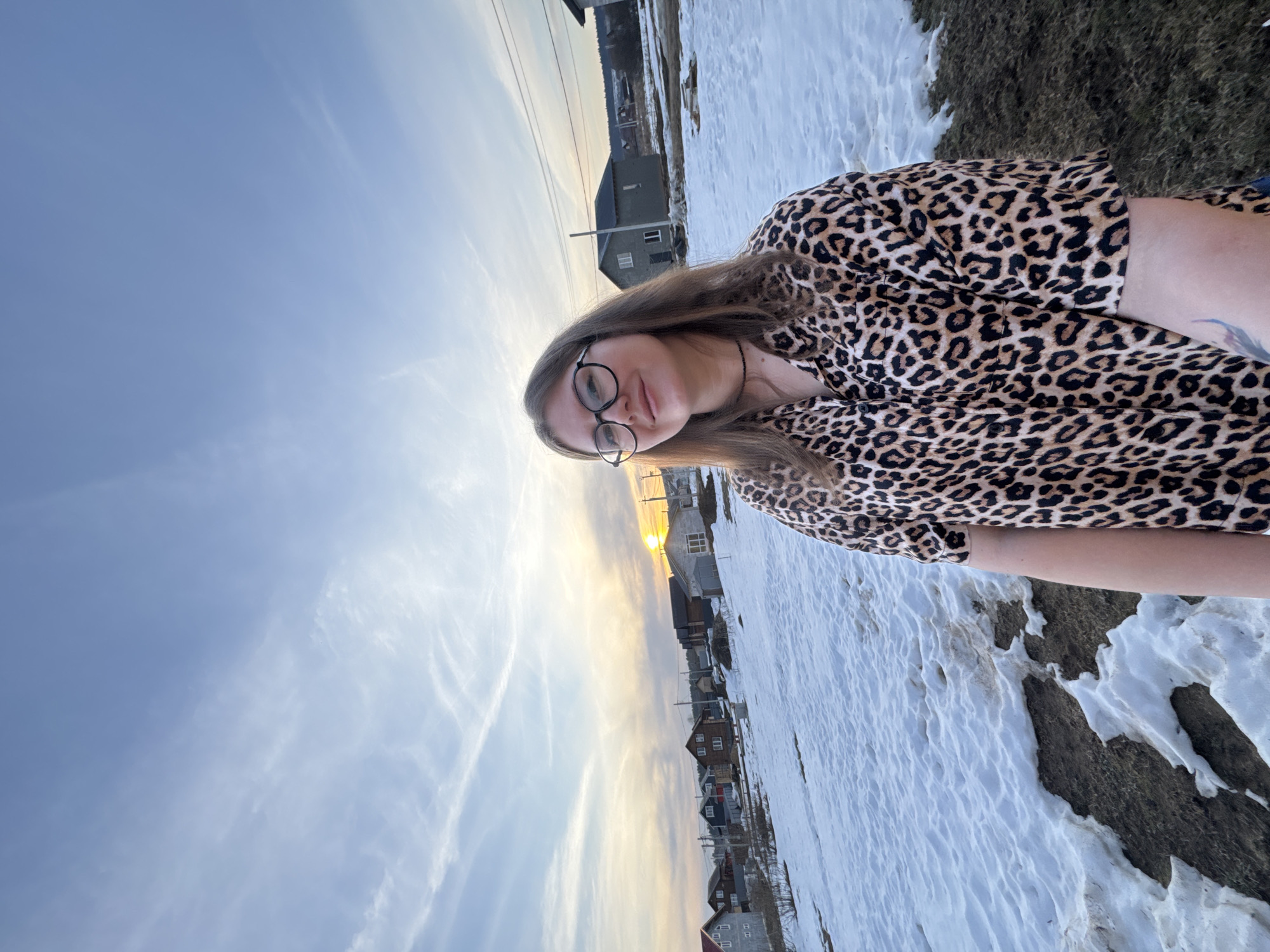 The image depicts a woman with glasses and long hair, wearing a leopard print top. She is standing outside against a backdrop of a sunset and houses. The location is in Gornyi (Dvurechenskoe S/P), Russia, as evidenced by the presence of characteristic Russian houses. There is snow visible on the ground. The time of day is likely late afternoon or early evening, given the setting sun.