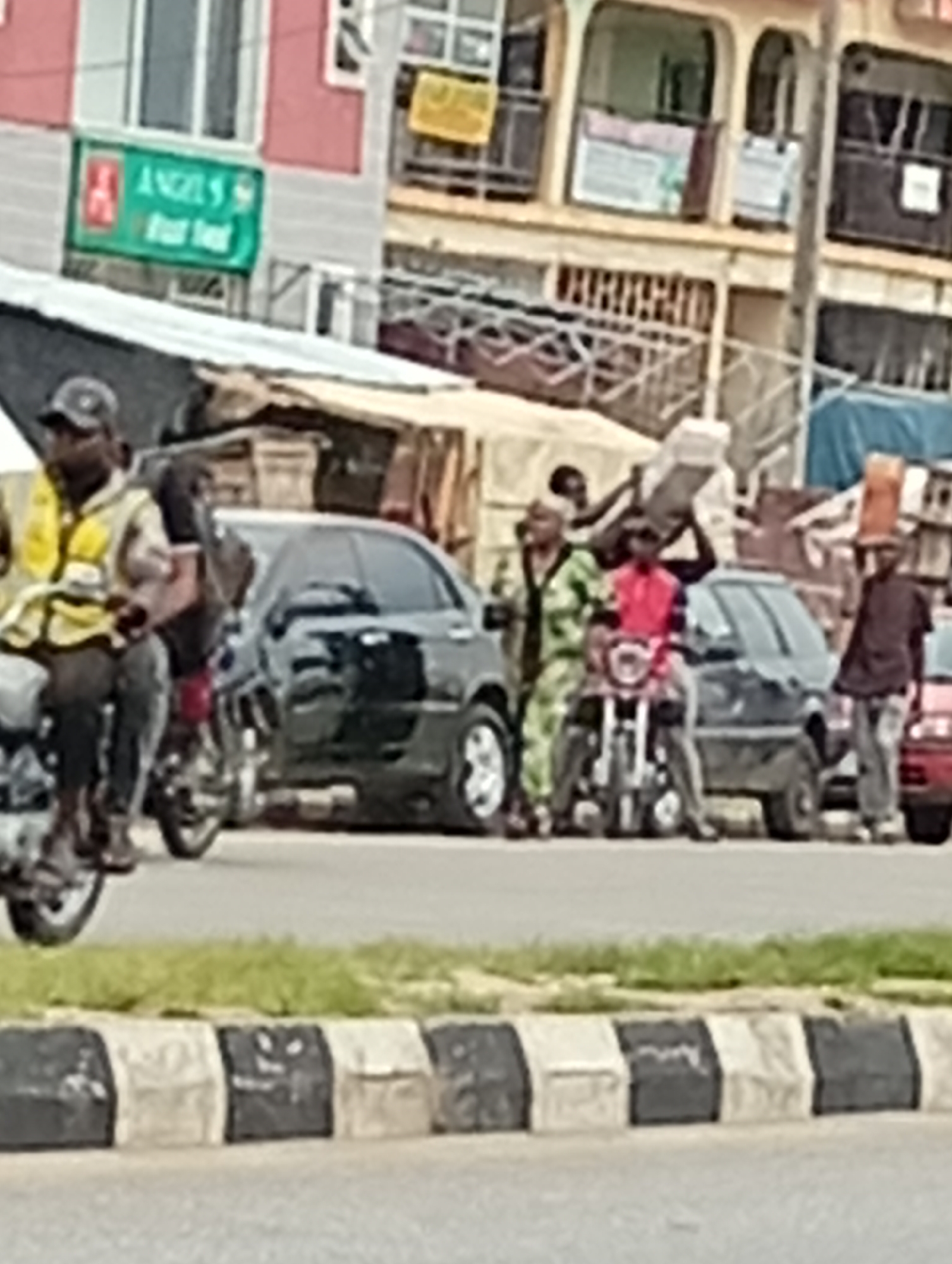 In an outdoor urban scene, a group of people are gathered near a street with several vehicles. On the left, a person wearing a yellow vest and a dark cap rides a motorcycle. To their right, a black sedan is parked. Further in the background, several people are seen in the middle of the street, some carrying large rectangular objects. One individual in a red shirt is seated on a motorcycle. To the right, a person in a dark shirt and jeans stands near a red car. Behind the people and vehicles, buildings with multiple stories and various signs are visible. The street has a curb with alternating black and white markings. Green grass and foliage line the side of the road. The overall image quality is low, with blurred details.