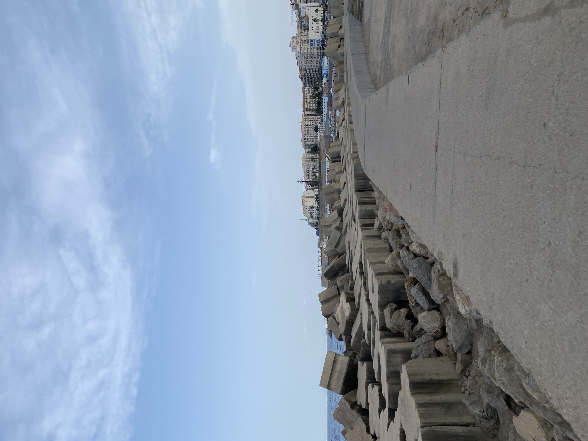 A concrete walkway curves along a coastline protected by large, irregular concrete blocks and smaller rocks. In the background, a city skyline with multi-story buildings, some featuring white and beige facades, stretches across the horizon under a pale blue sky with scattered clouds. The sea is visible to the left of the concrete structures, with a faint line of distant land or structures on the horizon. The scene is devoid of people and any apparent activity. The location is identified as Beau Fraisier, Algeria.