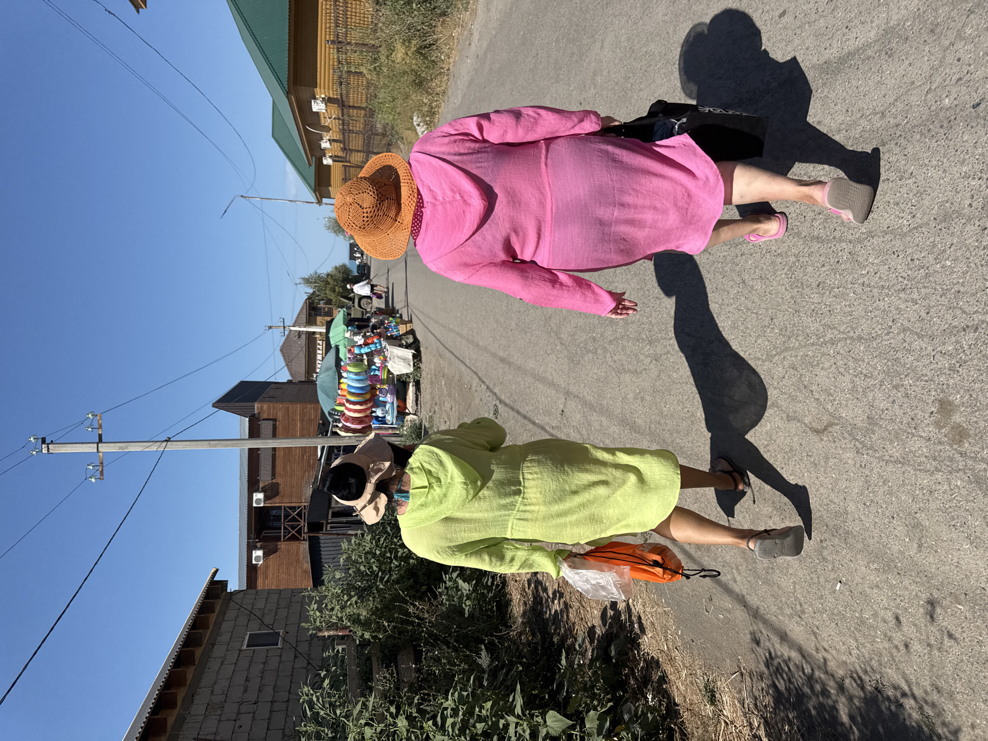 Two women are walking away from the camera down a paved street in Zharbulak, Kazakhstan. The woman on the left wears a lime green hooded robe and carries a clear plastic bag with an orange drawstring bag inside. The woman on the right wears a bright pink hooded robe and an orange crocheted hat, and carries a black tote bag. Both women are wearing sandals. The street is lined with buildings, including a brick building on the left and wooden buildings with green roofs on the right. In the distance, there are stalls with colorful merchandise, including inflatable rings. The sky is clear and blue. The sun casts long shadows on the street.