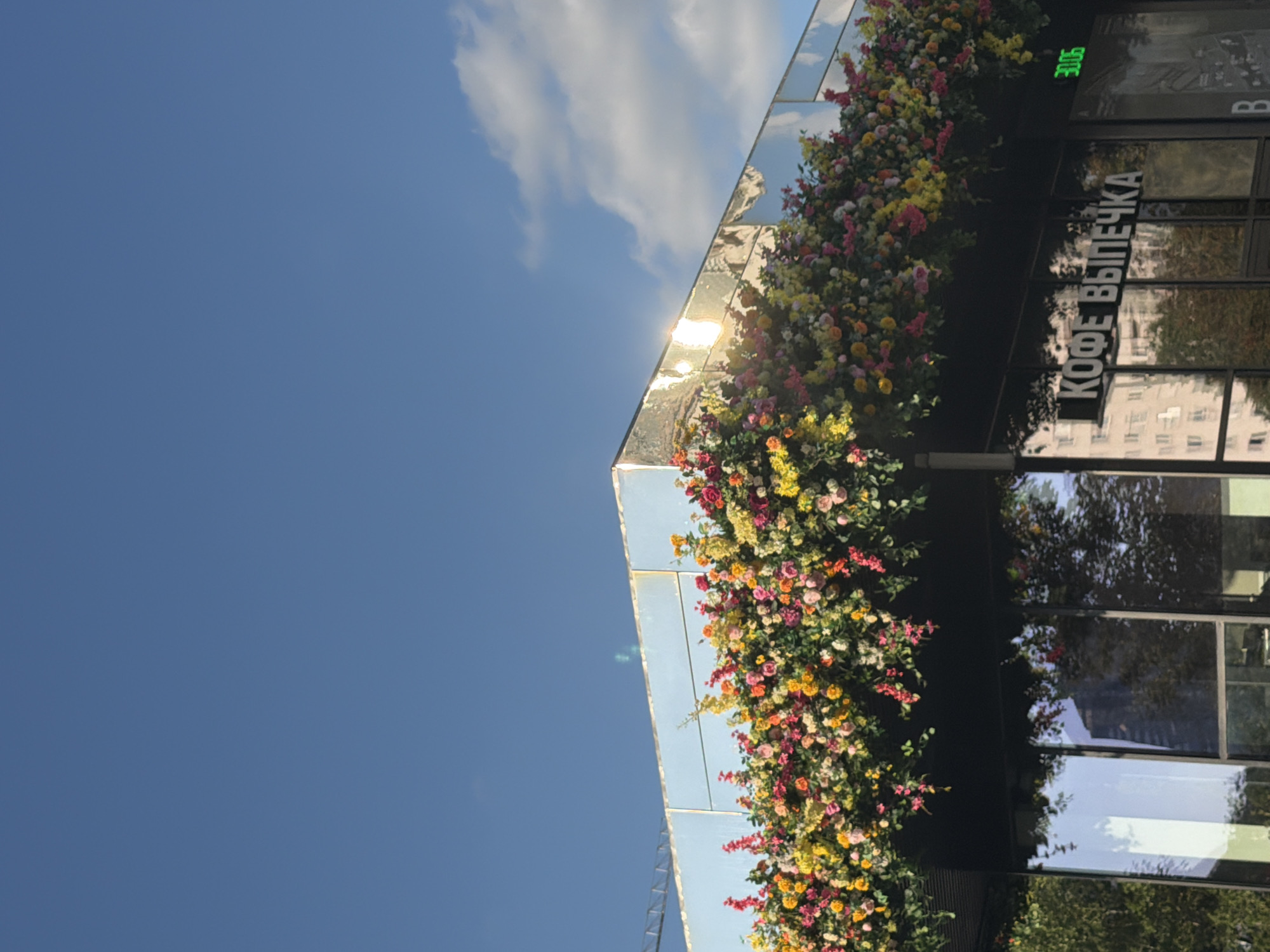 This image shows a close-up of a modern building with a mirrored facade and a vibrant floral arrangement adorning its corner. The bright blue sky with scattered white clouds suggests it is daytime, likely sunny due to the bright reflections on the building. The building's facade is clad in large, square mirrored panels, creating a striking visual effect that reflects the sky, clouds, and surrounding greenery.
A dense, colorful floral display cascades down the corner of the building, featuring a mix of pink, yellow, and orange flowers, interspersed with green foliage. The flowers appear to be a variety of small blossoms, creating a lush and festive appearance.
Visible on the dark surface of the building, to the right of the flowers, is white text that reads "КОФЕ ВЫПЕЧКА", which translates to "COFFEE BAKERY" in Russian. Above this text, a green digital display shows the number "3006". The reflection on the mirrored panels reveals the presence of trees and other buildings, indicating an urban or commercial setting. The overall impression is one of a stylish and well-maintained storefront, possibly a cafe, decorated for a special occasion or simply to enhance its aesthetic appeal.