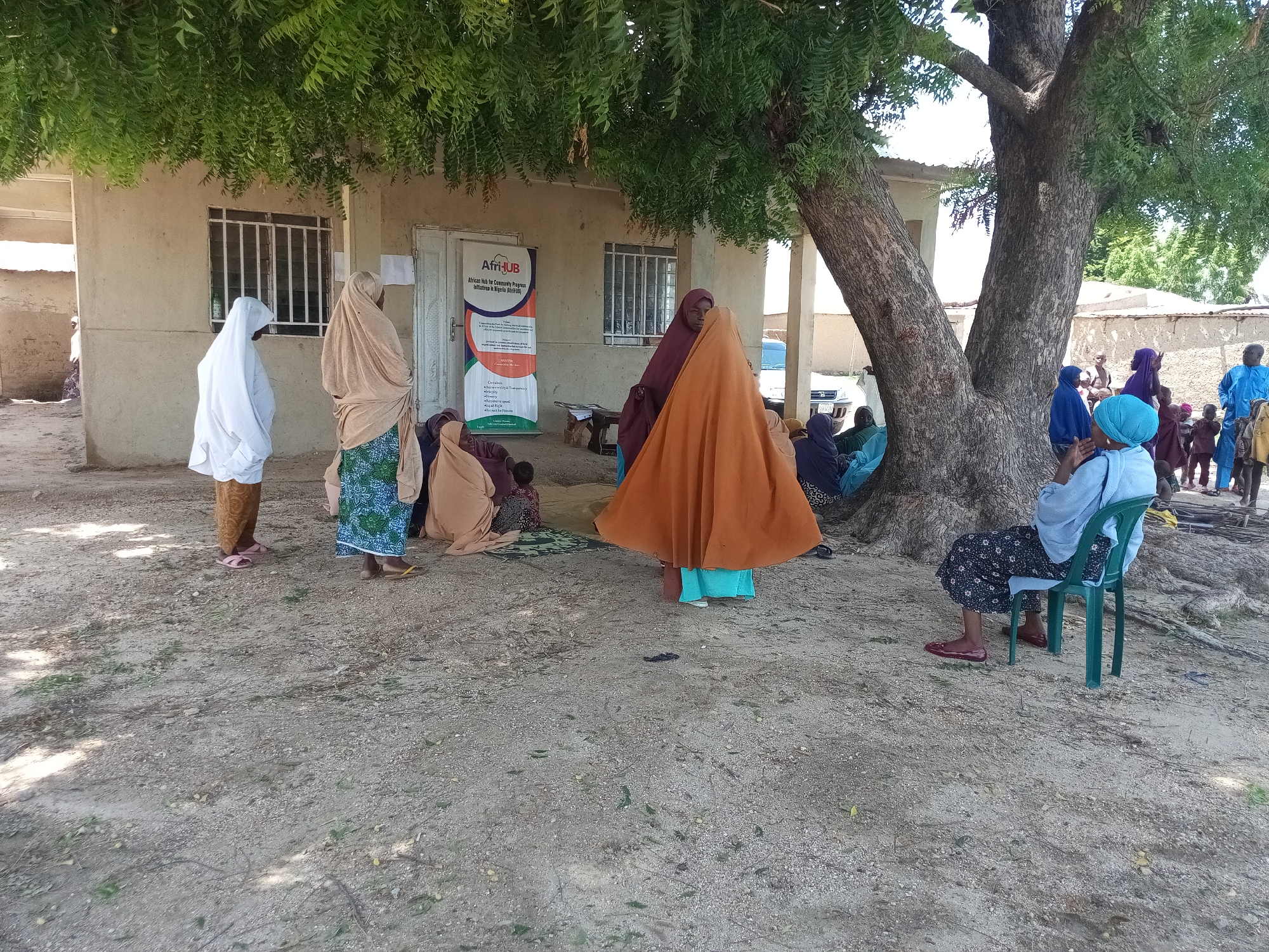 The image depicts an outdoor scene near a light-colored building, possibly a community center or clinic, with a large tree providing shade. A group of people, primarily women and children, are gathered. Several women are wearing hijabs and traditional clothing, with some sitting on the ground and others standing. A banner with text and logos, including "AfriHUB," is displayed on the side of the building. The ground is covered in sand and dirt, with some scattered leaves and debris. In the background, more buildings and greenery are visible, suggesting a rural or semi-urban environment. One woman is seated on a green plastic chair. The overall atmosphere appears to be one of a community gathering or waiting area.