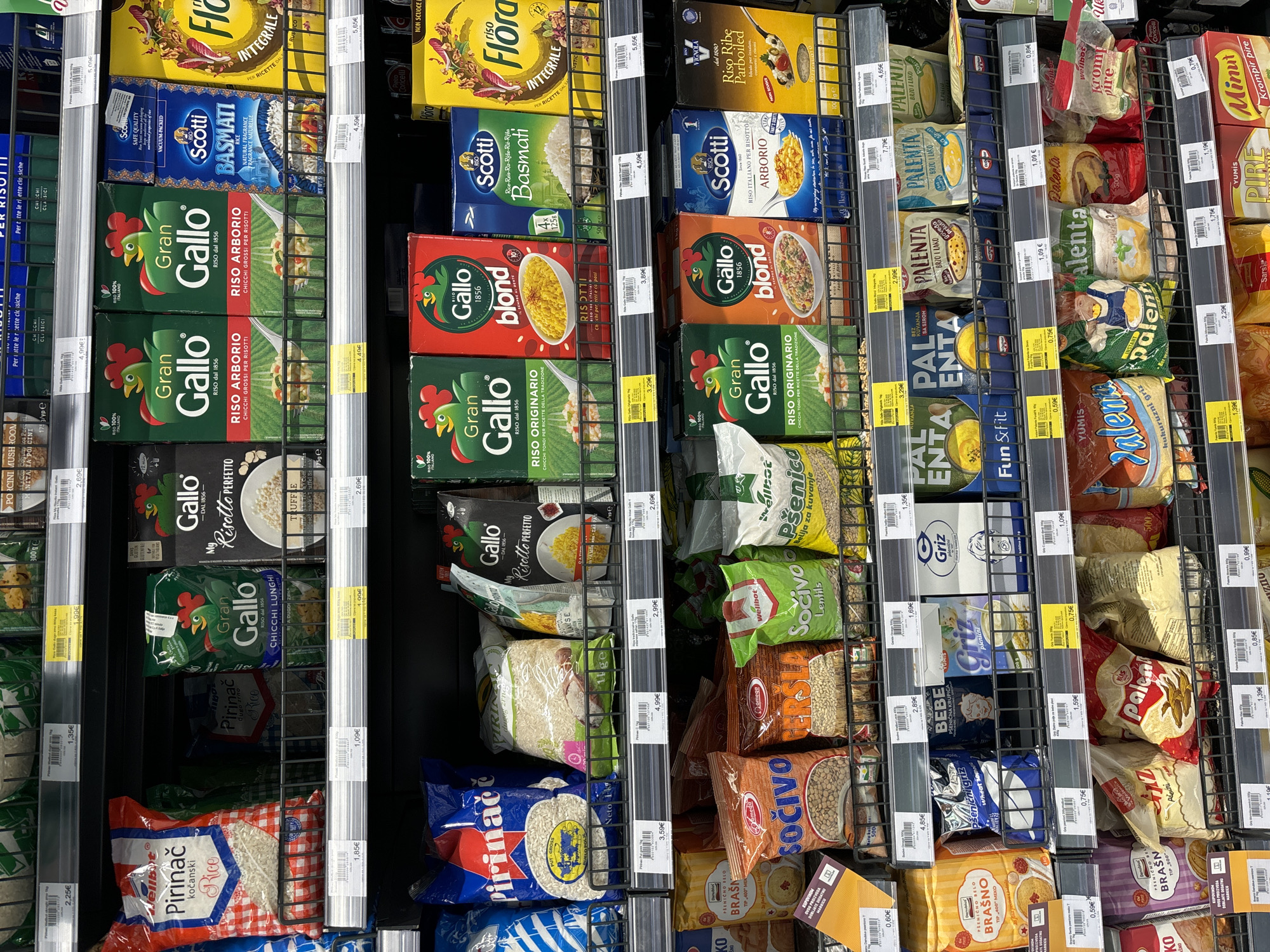 The image shows shelves of rice and other grain products in a supermarket. The shelves are well-stocked, with various brands and types of rice, including Arborio, Basmati, and integral rice. There are also bags of lentils, flour, and semolina. The products are neatly arranged, and the lighting is bright and even. The overall impression is one of a clean and organized supermarket.
The location is a supermarket in Budva, Montenegro. This is suggested by the presence of products with labels in Cyrillic and Latin script, common in the Balkan region, and the overall style of the supermarket which is typical of many in Eastern Europe. The specific type of store is likely a large supermarket or hypermarket given the extensive selection of products and the multi-tiered shelving system. There are no people visible in the image.