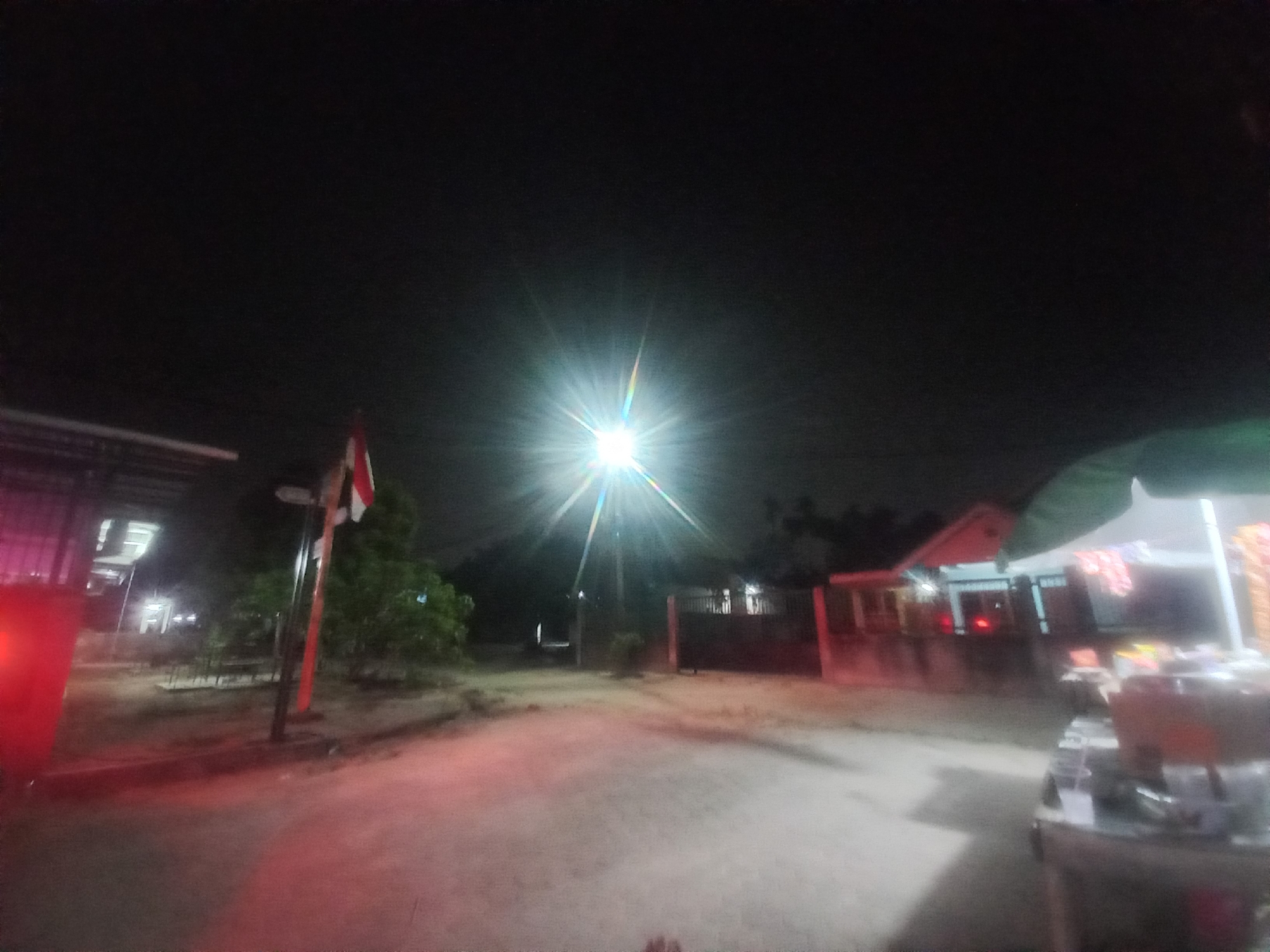 The image depicts an outdoor scene at night, likely in Kasang Pudak, Indonesia, illuminated by a bright overhead street lamp that creates a starburst effect. In the foreground, a dirt or gravel path is visible, with scattered debris. To the left, a red structure, possibly part of a building or vehicle, emits a red glow onto the ground. Further back, a flag with red and white stripes is attached to a pole, adjacent to a small tree. The middle ground shows a building with a covered entryway and some outdoor seating. To the right, a food stall or cart is present, with stacked containers, and what appear to be cups or glasses. The background is dark, with some indistinct structures and foliage. The overall impression is a dimly lit, possibly commercial or public area at night.