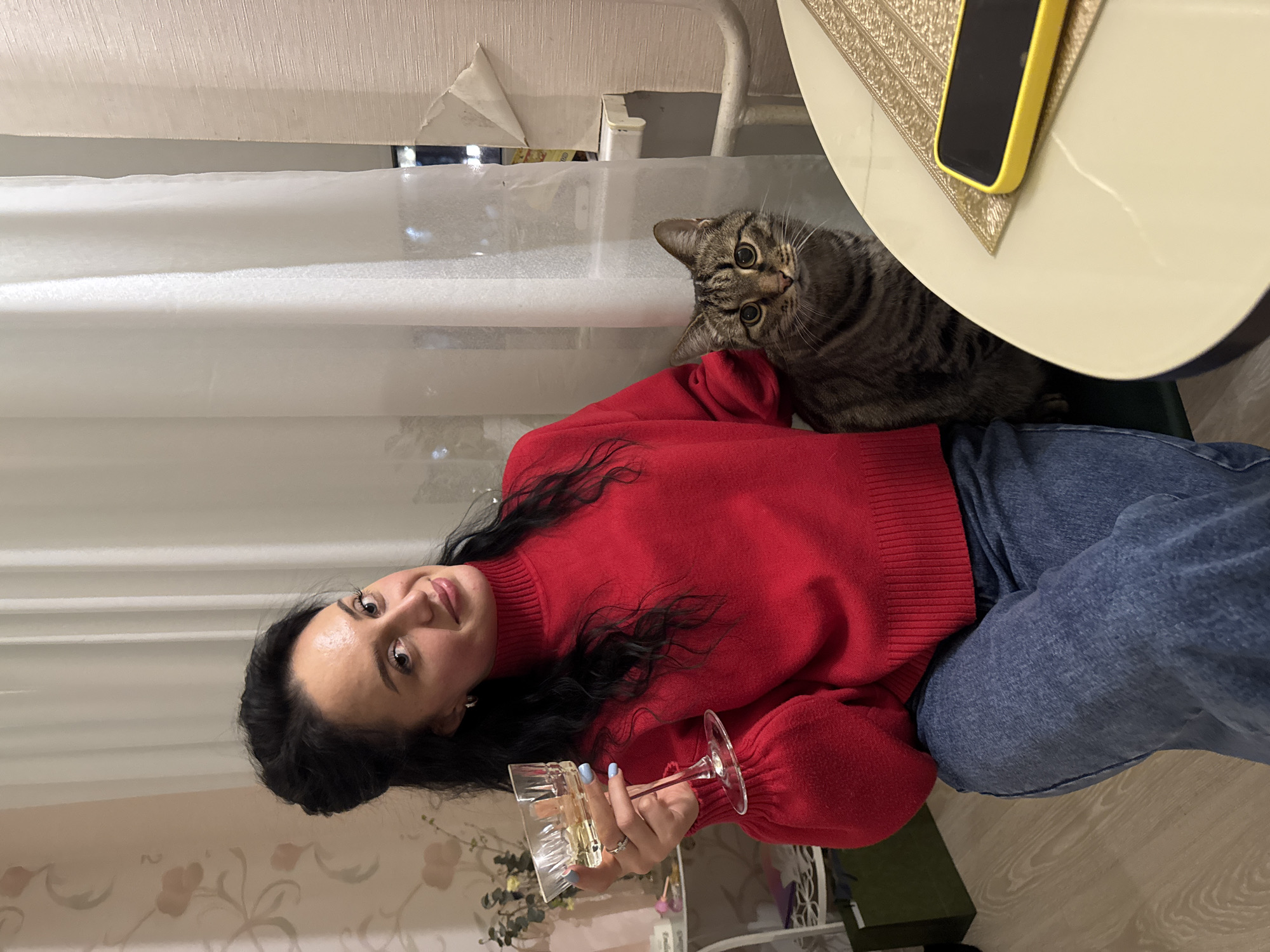 The scene is an interior residential space located in Chaikovsky, Russia. An adult female, with dark wavy hair and wearing a red turtleneck sweater and blue jeans, is seated, facing slightly right and looking directly at the camera with a subtle smile. Her right hand, featuring light blue nail polish and a ring, holds a stemmed glass containing a clear, light-yellow liquid. To her right, a domestic tabby cat with brown and black stripes and prominent eyes sits upright, also looking directly forward. Both subjects are positioned alongside a round, light-colored table, upon which a yellow-cased smartphone lies face down on a textured gold placemat. The background features light-colored walls; the left wall displays a faint floral pattern, while the right wall exhibits textured wallpaper with a visible tear or peel near a window. White, sheer curtains cover the window, behind which outdoor lights are visible, indicating an evening setting. A white radiator or pipe runs horizontally below the window. A light wood-grain floor is visible in the lower left, adjacent to a dark green bag. A white shelving unit with decorative items is partially visible on the left.