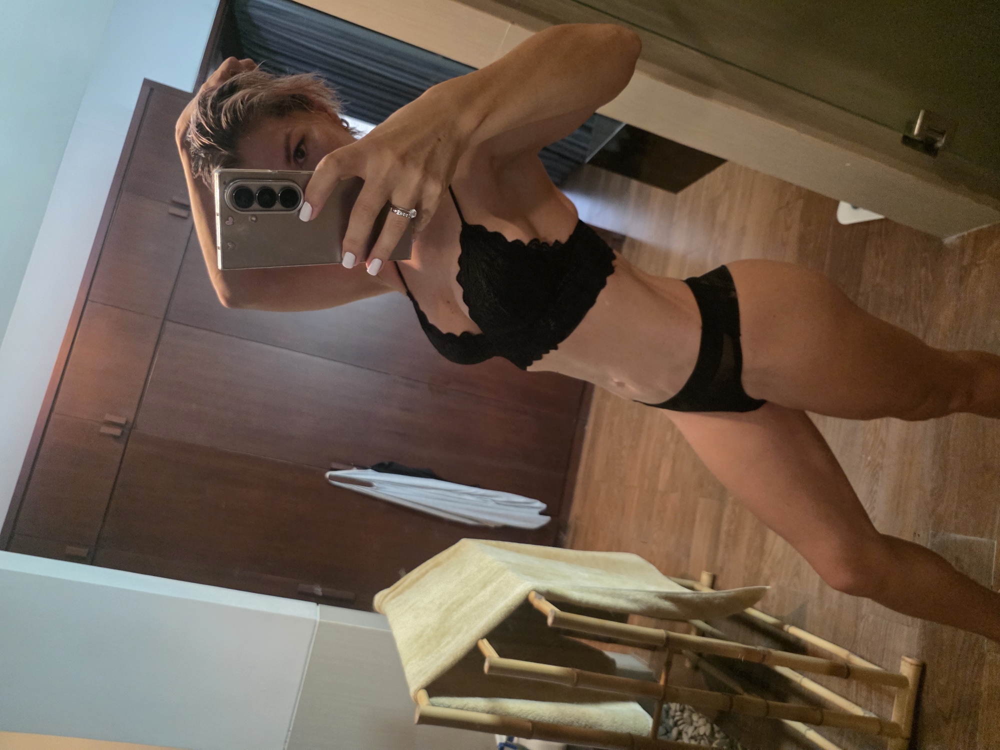 A woman is taking a mirror selfie in what appears to be a bedroom or dressing area. She is wearing black lace lingerie and has her hair styled short and tousled. Her left arm is raised behind her head, and her right hand is holding her phone. The background includes wooden wardrobes, a towel rack with a tan towel, and a wooden floor.