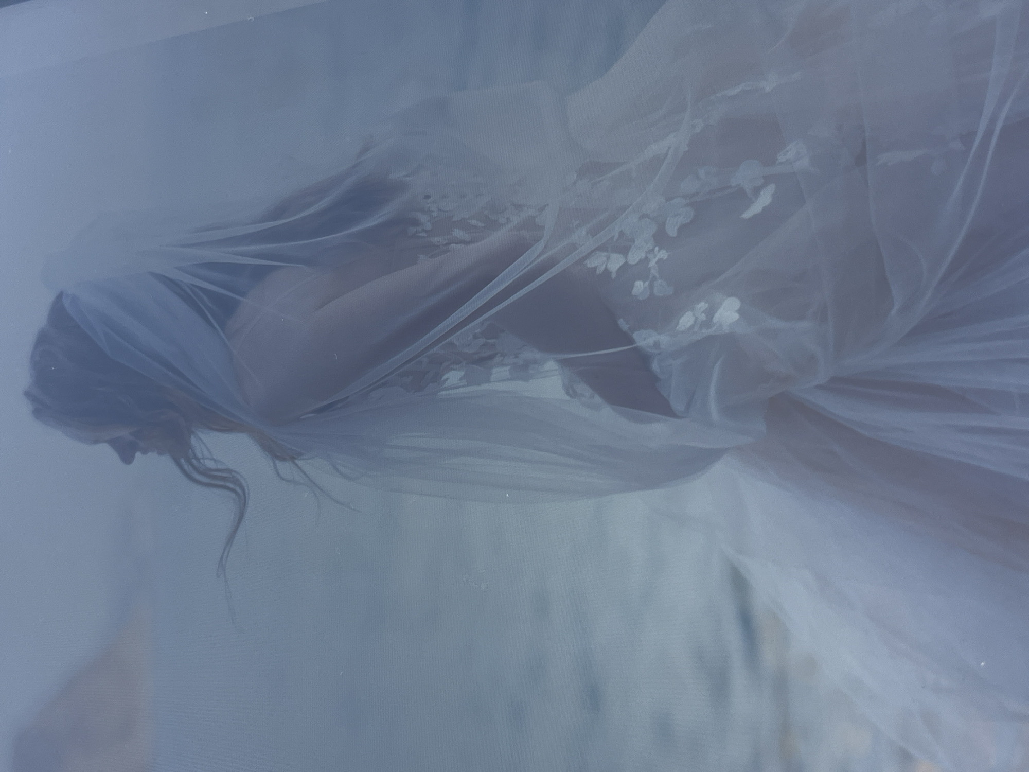 A veiled woman is depicted in this photograph. Her face is partially obscured by the veil, and part of her hair is visible. The veil also covers her shoulder and part of a white dress, which features floral embroidery. The background is muted. The location is unknown.