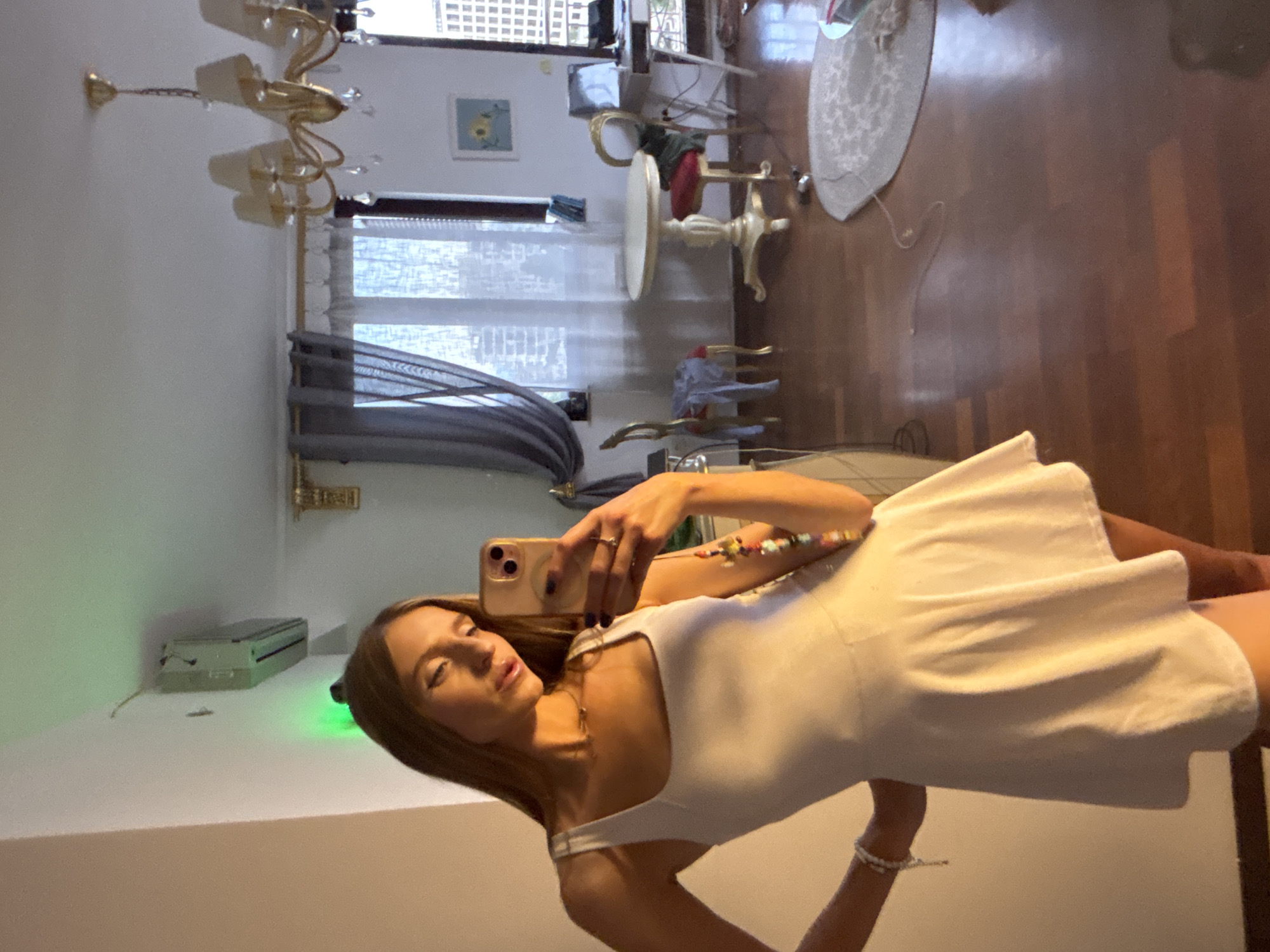 A young woman with long blonde hair takes a selfie in a room. She is wearing a white sleeveless dress and multiple bracelets. She is holding a smartphone in her right hand and posing with her left hand on her hip. The room appears to be a living room with wooden floors, a round table with chairs, a chandelier, and a window with curtains. The woman is standing near a wall with an air conditioning unit and a green light. The image appears to be taken during the day.