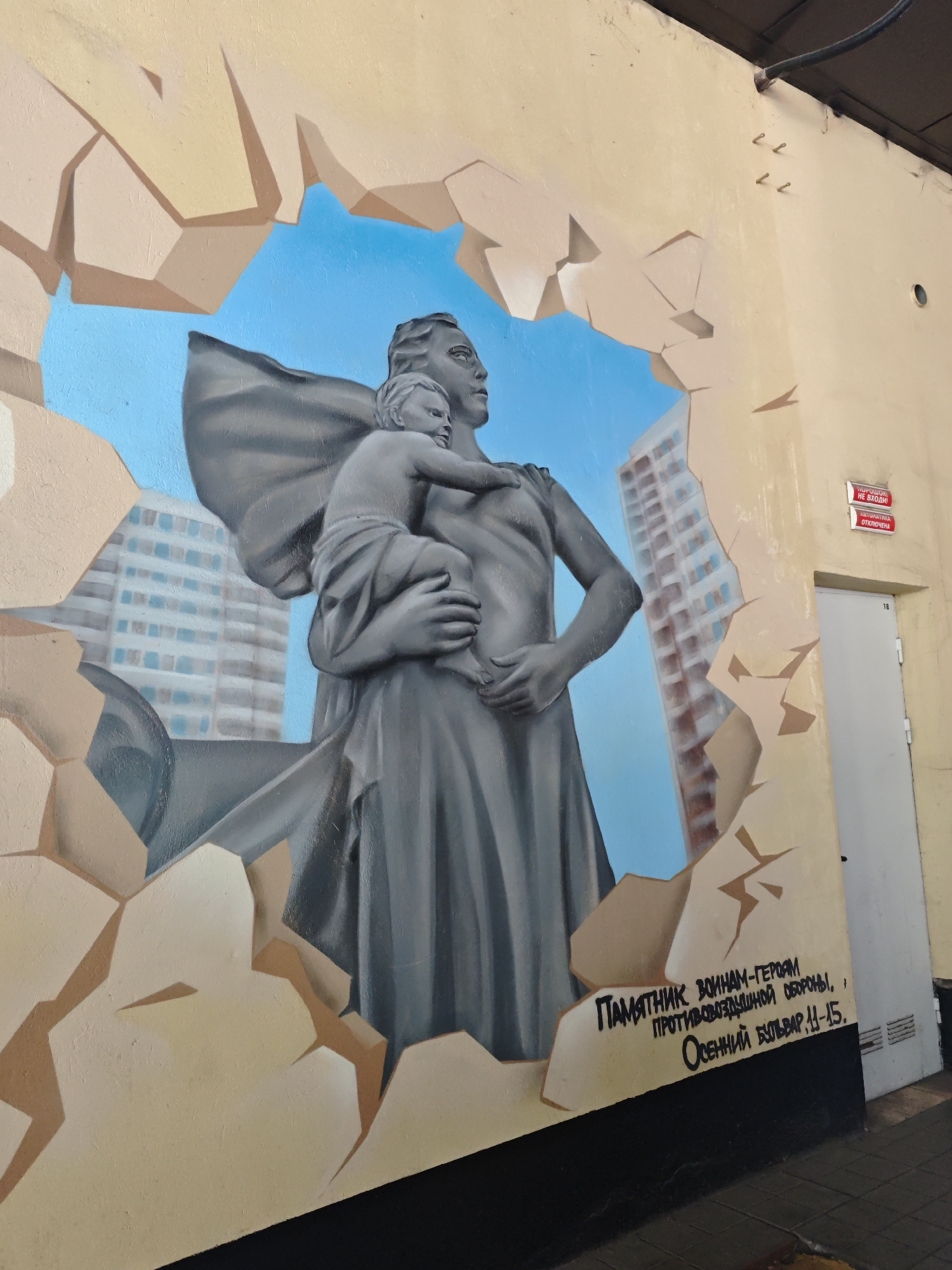 The image shows a mural painted on a light-colored wall, depicting a statue of a woman holding a child. The statue is rendered in grey tones and is positioned as if breaking through a cracked, light beige surface. Behind the statue, a blue sky is visible with stylized buildings. The text painted on the mural, in black Cyrillic script, reads "ПАМЯТНИК ВОИНАМ-ГЕРОЯМ ПРОТИВОВОЗДУШНОЙ ОБОРОНЫ, ОСЕННИЙ БУЛЬВАР, 11-15." This translates to "MONUMENT TO THE HERO-WARRIORS OF AIR DEFENSE, OLSENY BOULEVARD, 11-15." The mural is located on a building with a white door and two red signs above it. One sign reads "ПОЛЬЗОВАНЕ НЕ ВХОДИ!" (DO NOT ENTER!) and the other states "АВТОМАТИКА ОТКЛЮЧЕНА" (AUTOMATICS ARE DISABLED). The location is identified as Osenny Boulevard, building 11-15, in Moscow, Russia.