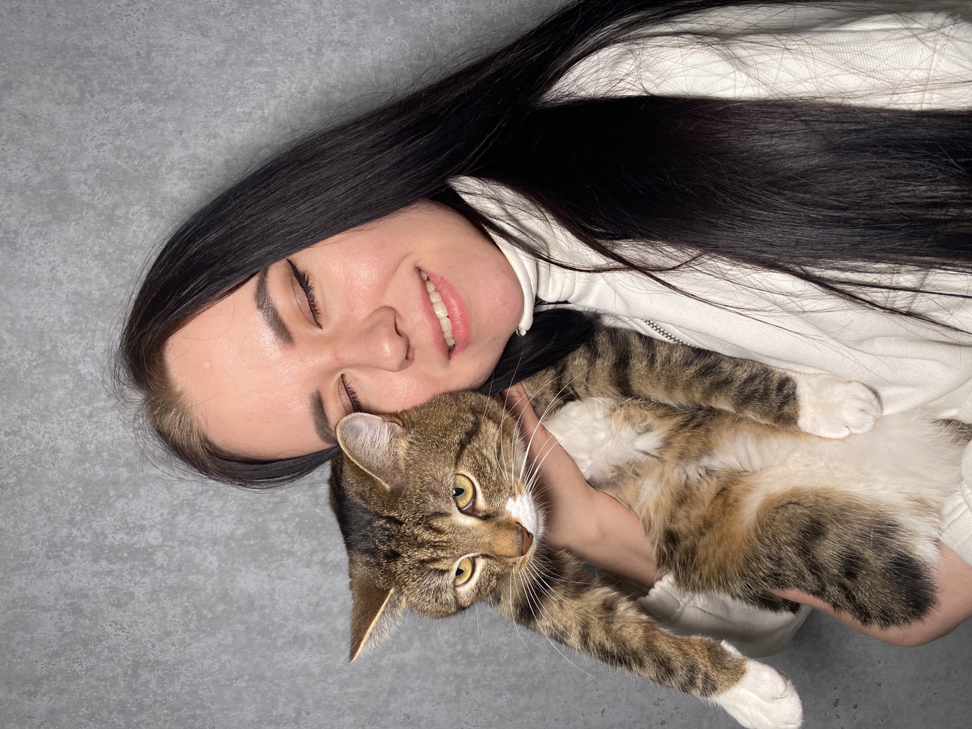A woman with long, dark hair and a white jacket is lying on a gray surface, holding a tabby cat. The woman is smiling with her eyes closed. The cat is looking directly at the viewer. The location is not specified.