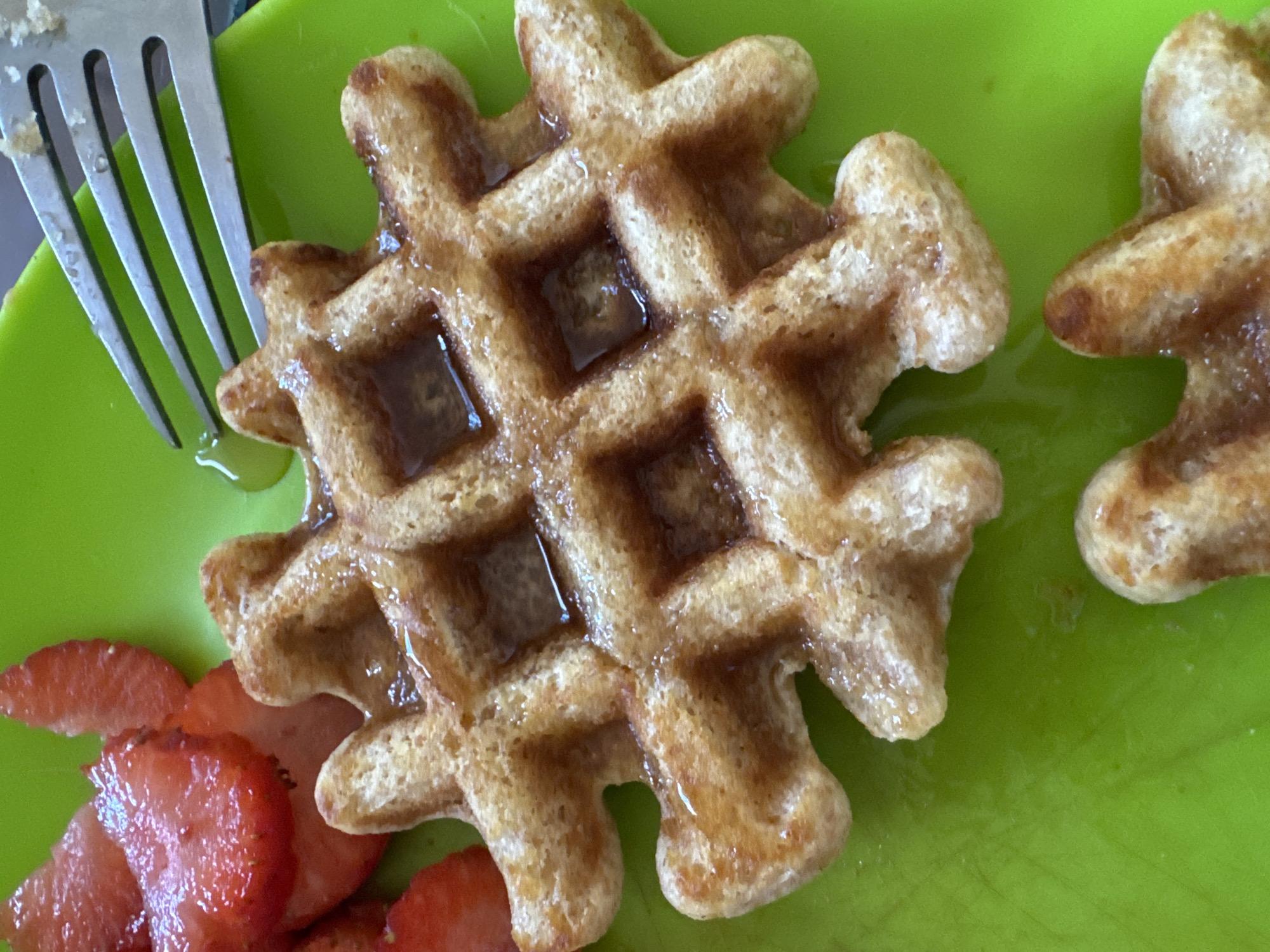 A close-up shot depicts a snowflake-shaped waffle topped with syrup, positioned on a bright green plate alongside sliced strawberries and a silver fork. Another similar waffle is partially visible at the edge of the frame. The specific location in Oaxaca City, Mexico is not discernible from the image.
