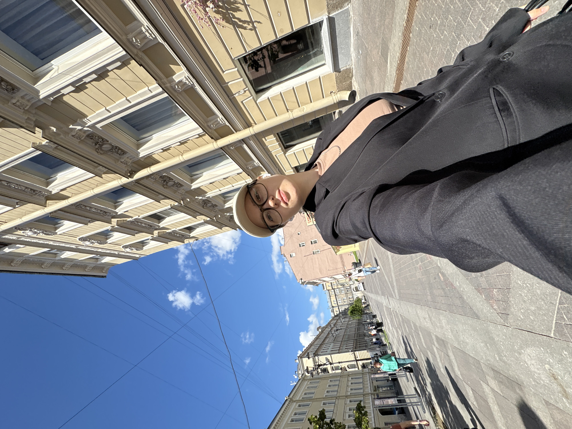 The image shows a woman taking a selfie on a sunny day on Nevsky Prospekt in Saint Petersburg, Russia. She is wearing a black blazer over a beige top and a white baseball cap, with glasses. The background features the distinctive architecture of Nevsky Prospekt, with its ornate yellow and beige buildings, and a clear blue sky with scattered clouds. The street is paved with granite tiles and there are several people walking along the sidewalk, as well as lampposts and trees. The perspective of the selfie is wide-angle, with the woman's arm extended to capture herself and the street scene.