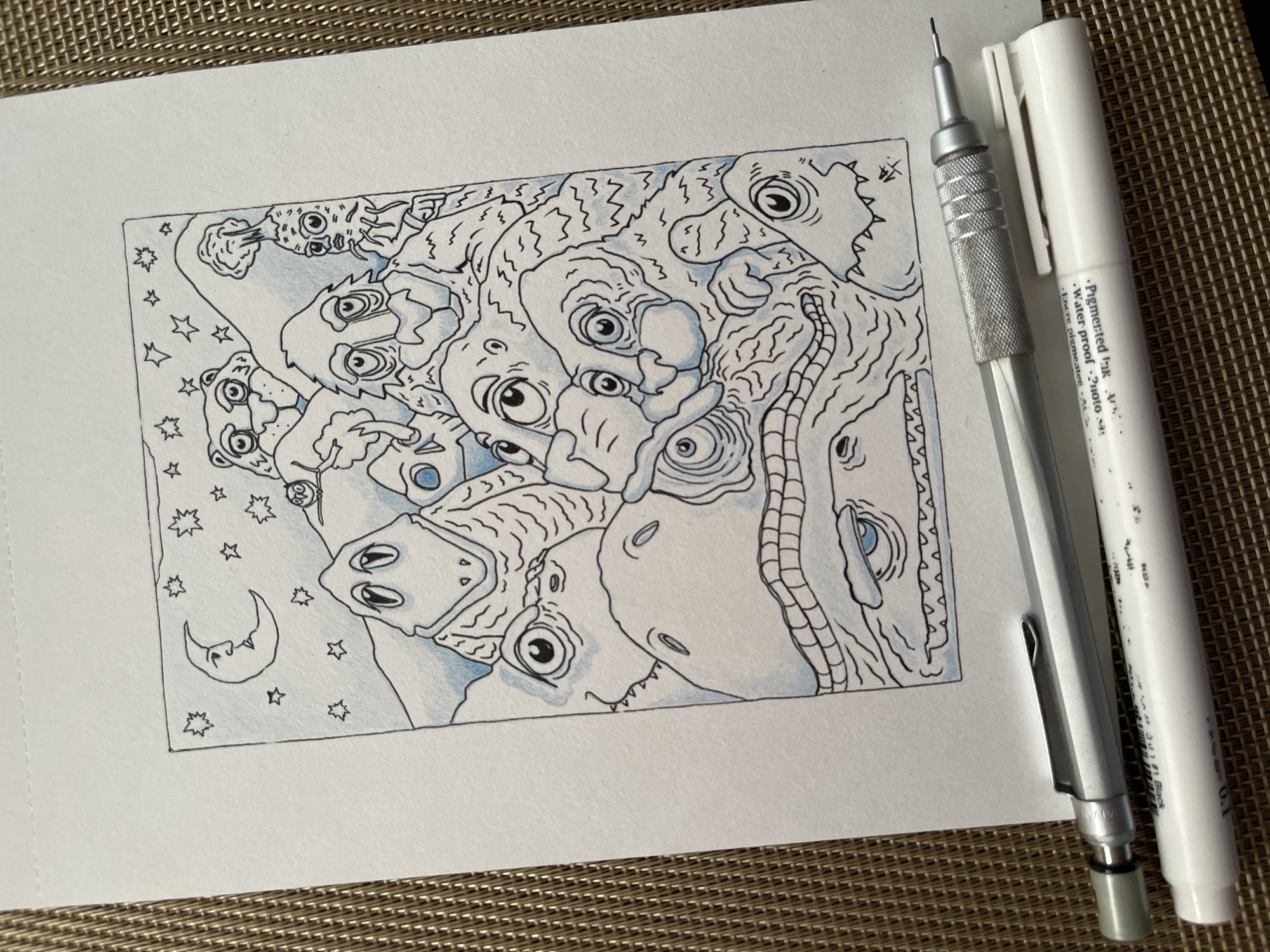 A drawing on paper depicts a group of whimsical, alien-like creatures in a stylized landscape. The creatures have large, expressive eyes and varied textures, with some appearing scaly and others fuzzy. They are clustered together, suggesting a community or a shared experience. The background includes a crescent moon with a face, and scattered stars, set against rolling hills or mountains. The artwork is rendered in black ink with subtle blue watercolor washes, creating a moody and dreamlike atmosphere. The drawing is framed by a thin black border. Two drawing instruments, a mechanical pencil and a white pen, lie in the foreground, positioned across the bottom edge of the paper. The paper itself rests on a textured, woven surface, possibly a placemat or mat, with a brown and beige color palette. The scene is not indicative of any specific location in the city of Krasnoyarsk, Russia.