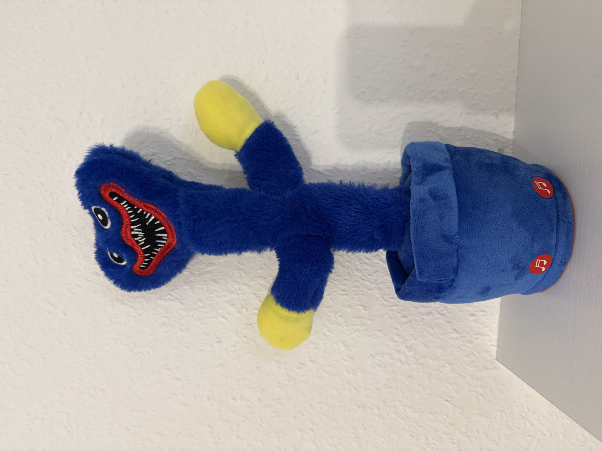A plush toy resembling the character Huggy Wuggy from the game Poppy Playtime stands in a blue pot on a white shelf against a textured white wall. The toy is predominantly blue with a large, toothy grin, red lips, and black and white eyes. It has yellow-tipped hands. The blue pot has two small red circular buttons with musical notes on them. This scene is located in Velbert, Germany.