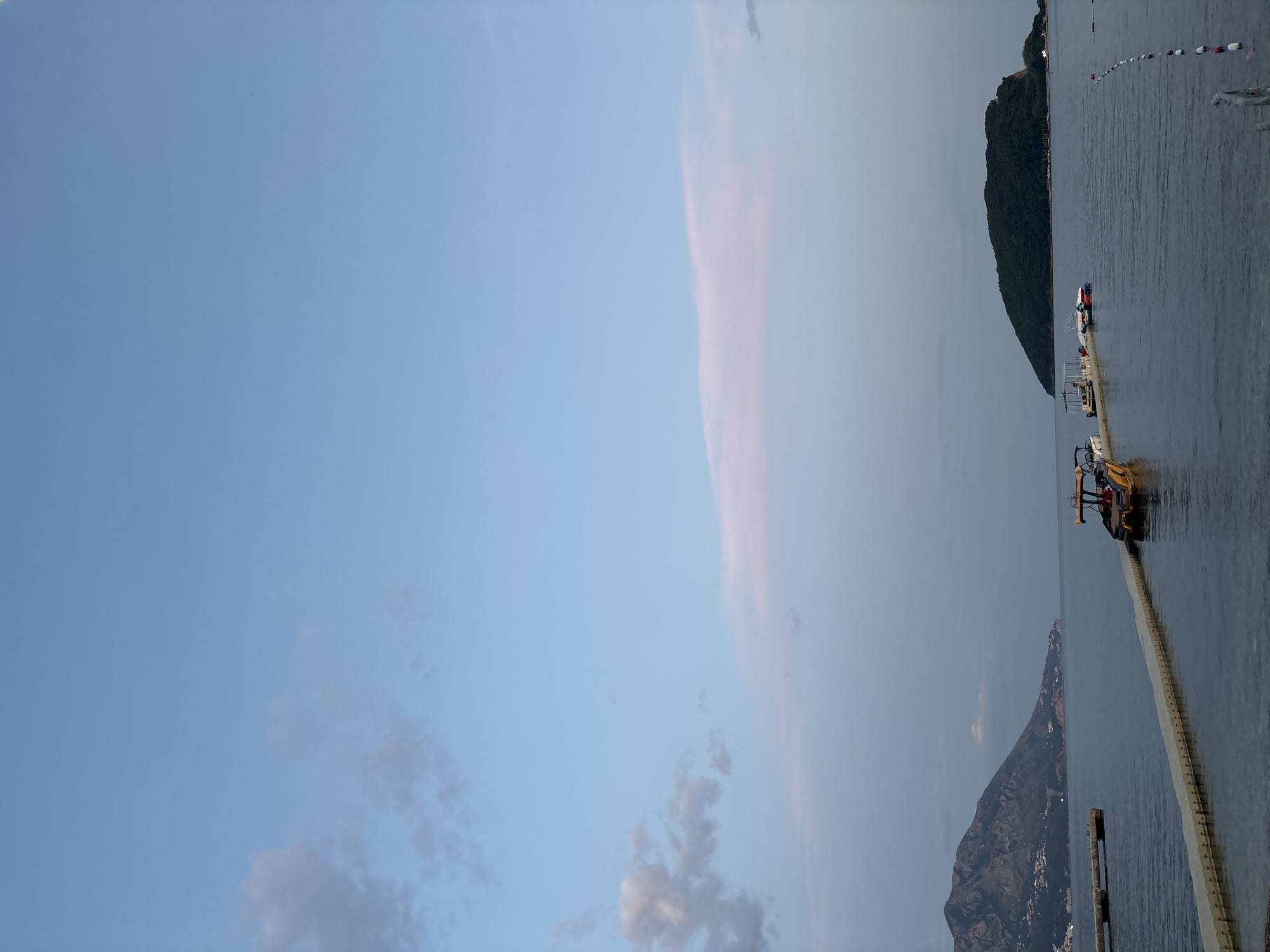 The image depicts a coastal scene, likely in Budva, Montenegro, given the landscape. The sky is predominantly blue with some cloud cover, transitioning to a hazy, lighter shade near the horizon. A body of water stretches across the right side of the image, possibly the Adriatic Sea. A green, mountainous landmass is visible along the shoreline. There's a long breakwater or similar structure extending into the water, with a piece of yellow machinery, such as a small excavator, situated on it. Faint buildings are visible along the edge of another land mass. A string of buoys, possibly marking a boundary for swimmers, is noticeable in the distance. The overall environment suggests a coastal construction or maintenance activity is taking place.